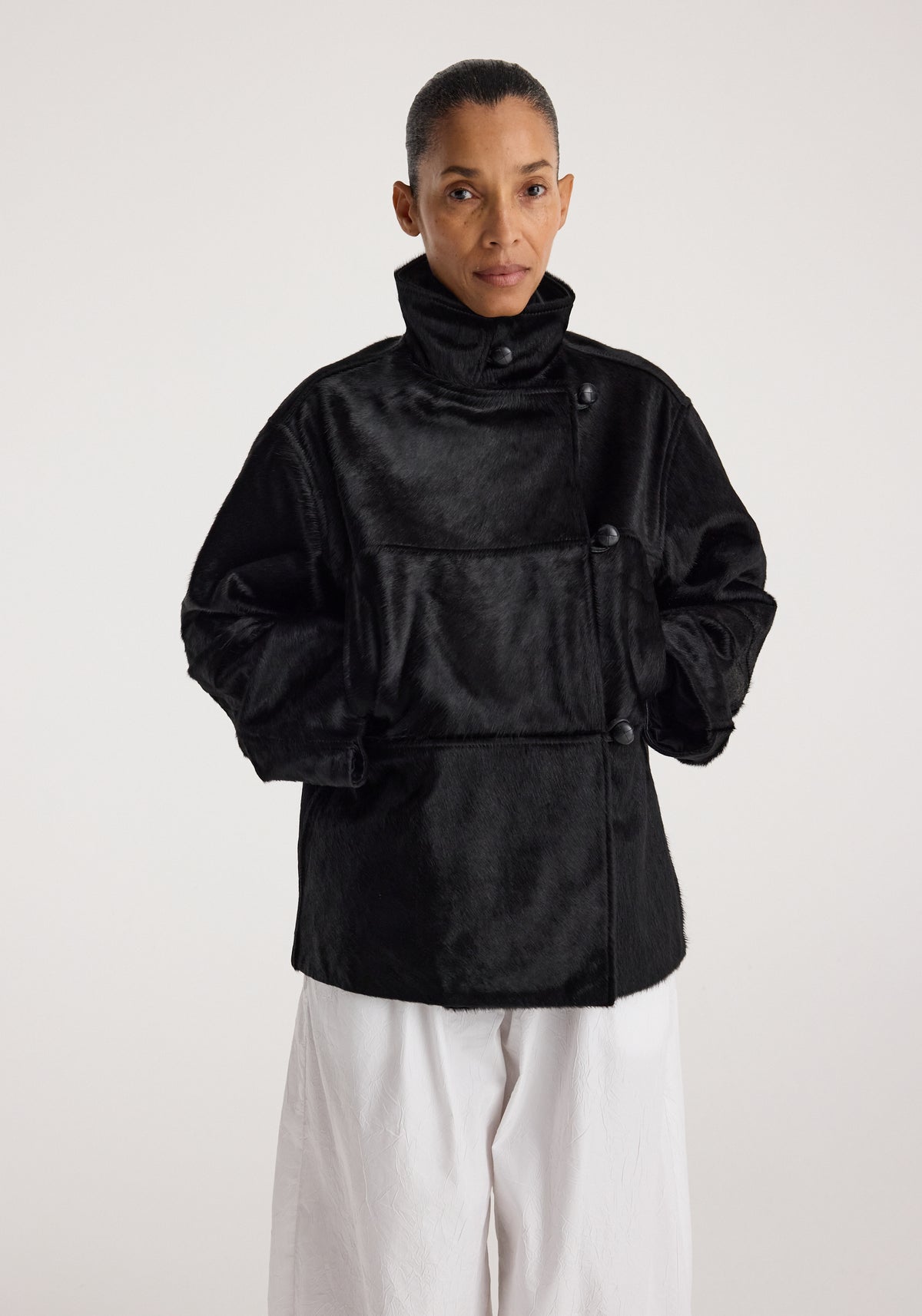 Woman wearing a black ponyhair jacket | black with a high collar and button closure, paired with loose white pants, standing against a plain background.