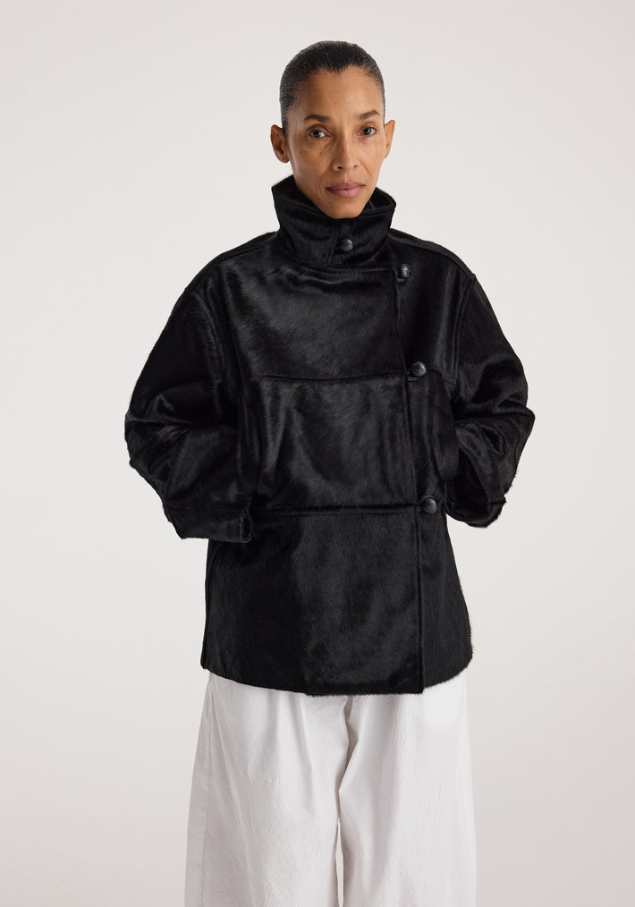Woman wearing a black ponyhair jacket | black with a high collar and button closure, paired with loose white pants, standing against a plain background.