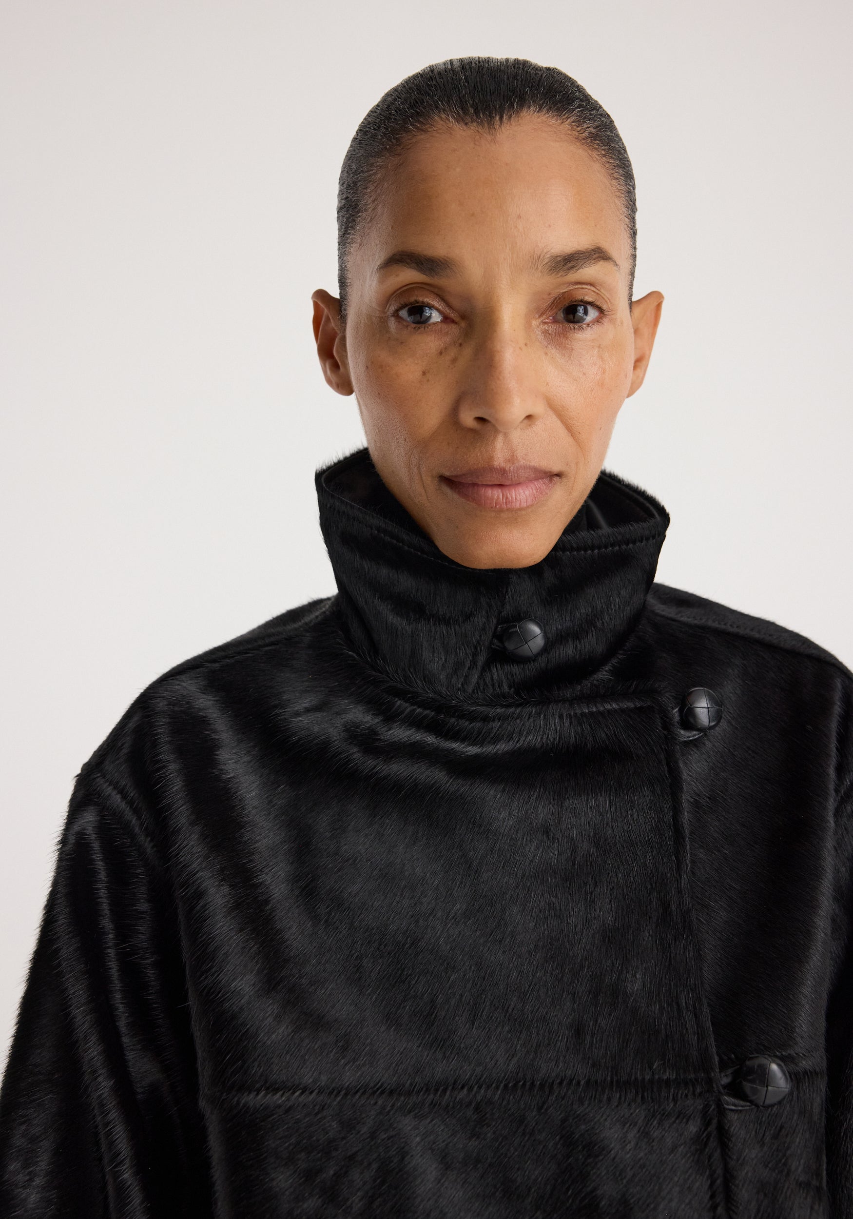 Close-up of a person wearing a high-collar Black ponyhair jacket | black with textured, shiny fabric and large buttons against a plain light background.