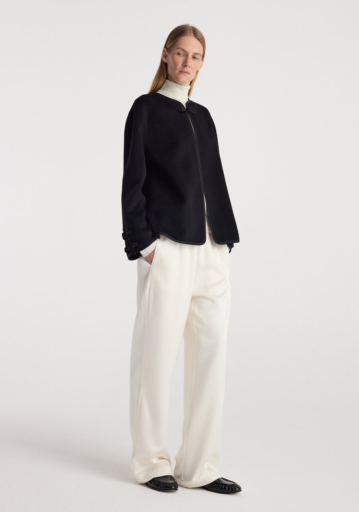 Woman wearing a black Double-faced pankou jacket | black with a zip front, paired with loose white pants and black shoes, standing against a plain white background.