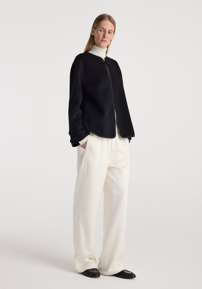 Woman wearing a black Double-faced pankou jacket | black with a zip front, paired with loose white pants and black shoes, standing against a plain white background.