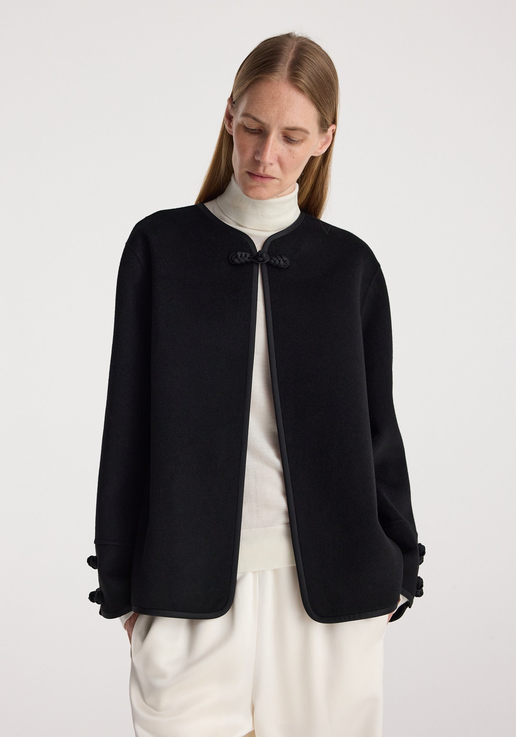 Woman wearing a black Double-faced pankou jacket | black over a white turtleneck and loose white pants, standing against a plain light background.