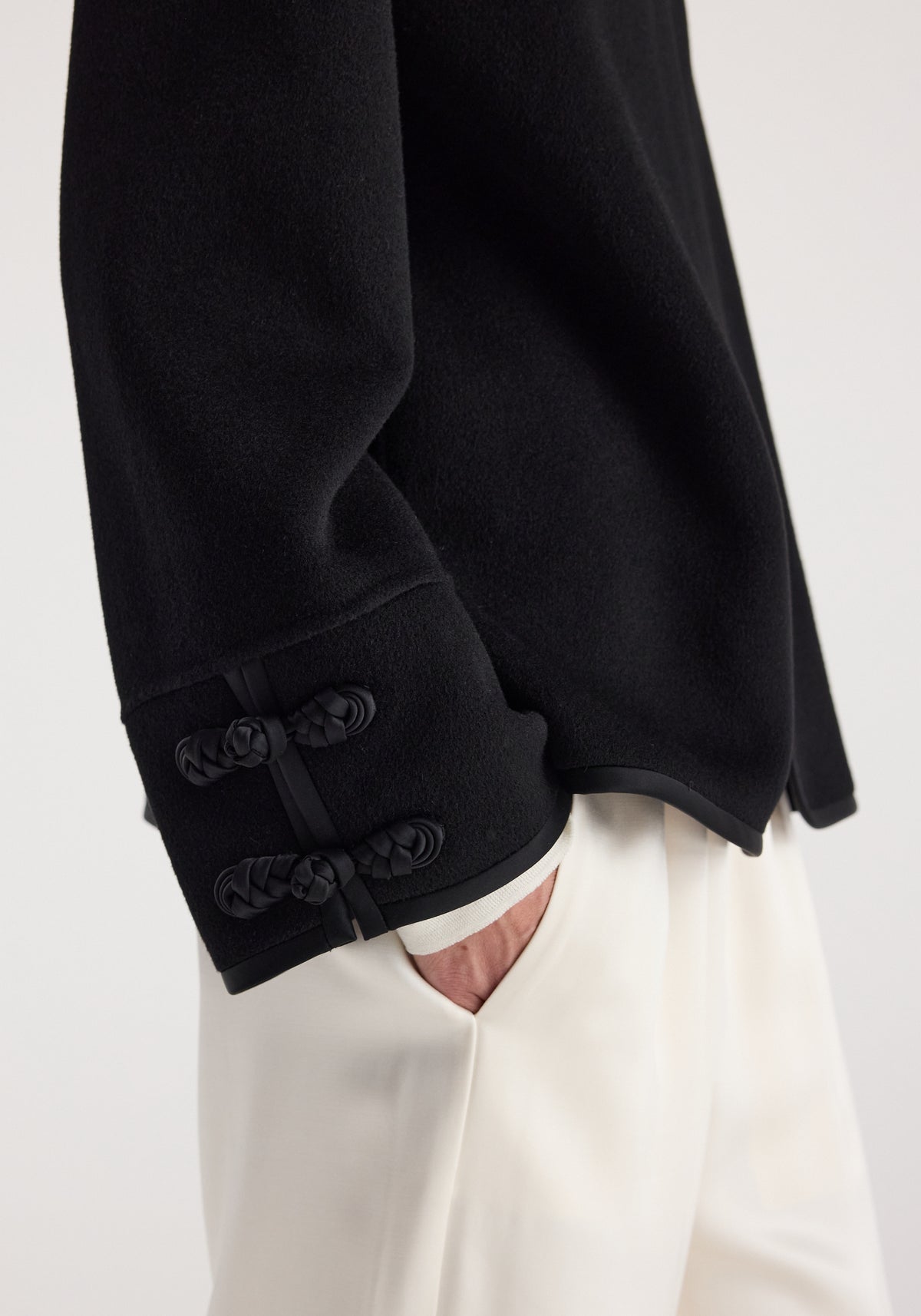 Close-up of sleeve detail on a Double-faced pankou jacket | black featuring intricate knot button closures, worn with white pants and a partially visible hand in pocket.