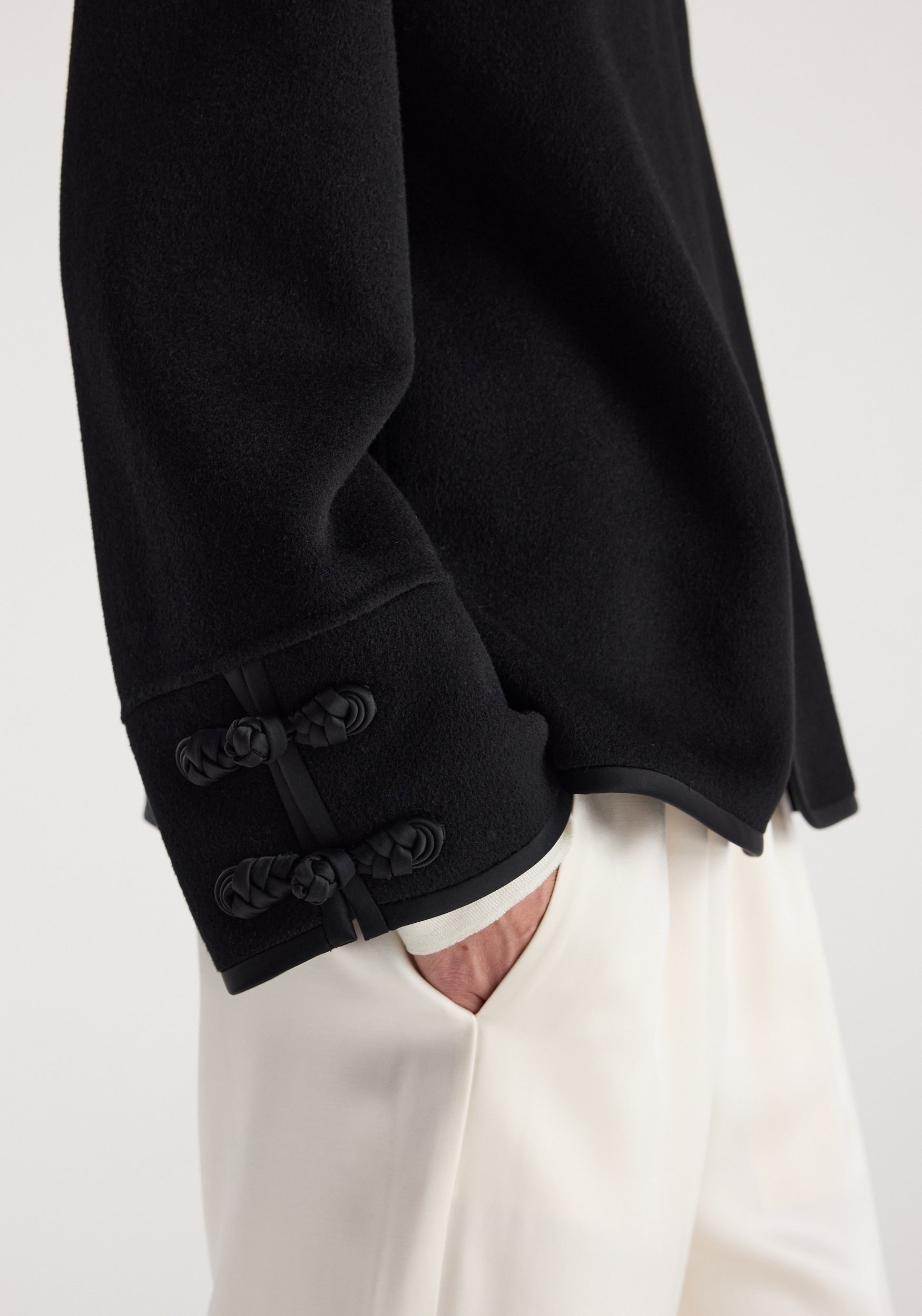 Close-up of sleeve detail on a Double-faced pankou jacket | black featuring intricate knot button closures, worn with white pants and a partially visible hand in pocket.