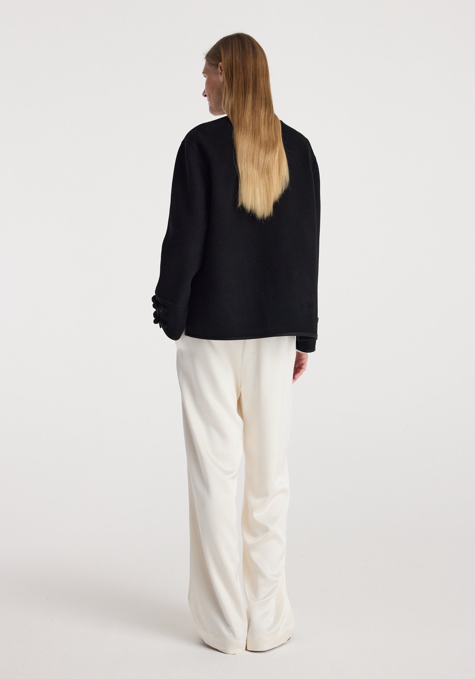 Woman seen from behind wearing a black Double-faced pankou jacket | black with detailed sleeves, paired with loose white pants against a plain background.