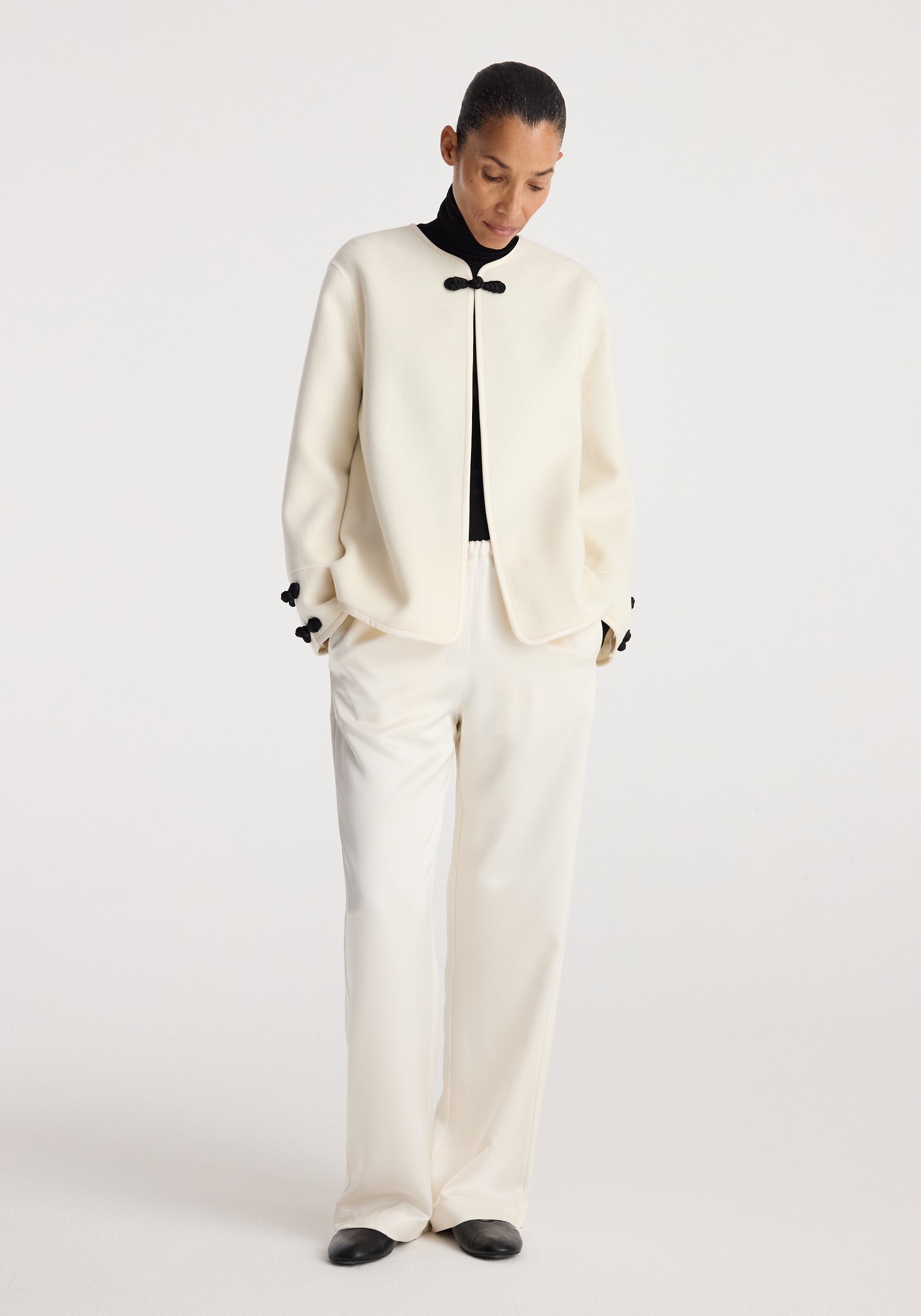 Woman wearing an off white Double-faced pankou jacket | off white with black decorative clasps, paired with matching wide-leg pants and black shoes against a plain background.