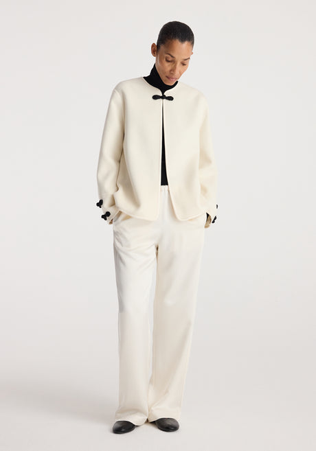 Woman wearing an off white Double-faced pankou jacket | off white with black decorative clasps, paired with matching wide-leg pants and black shoes against a plain background.