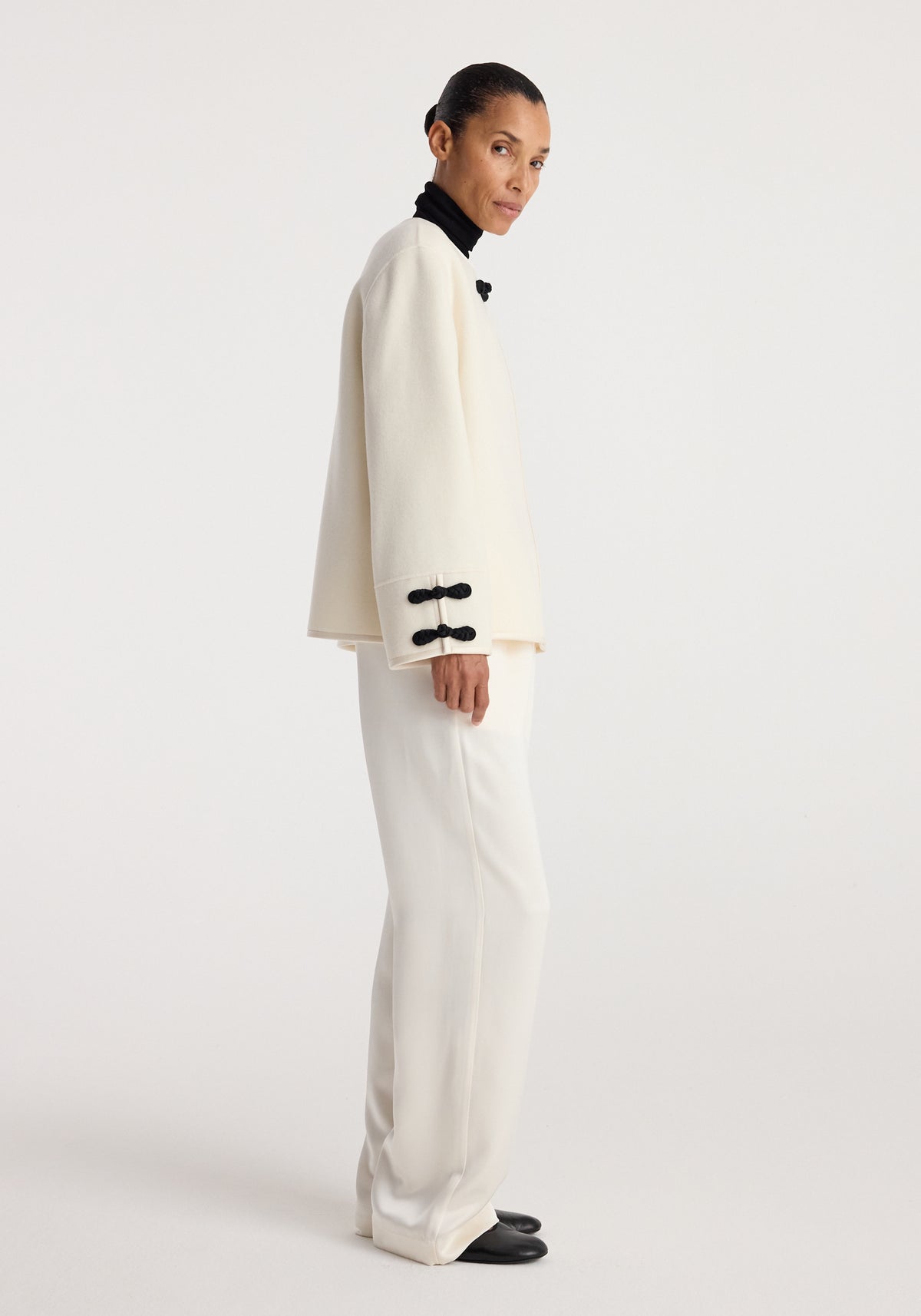 A model wearing a Double-faced pankou jacket | off white with black pankou button details on the cuffs and collar, paired with matching off-white pants and black shoes.