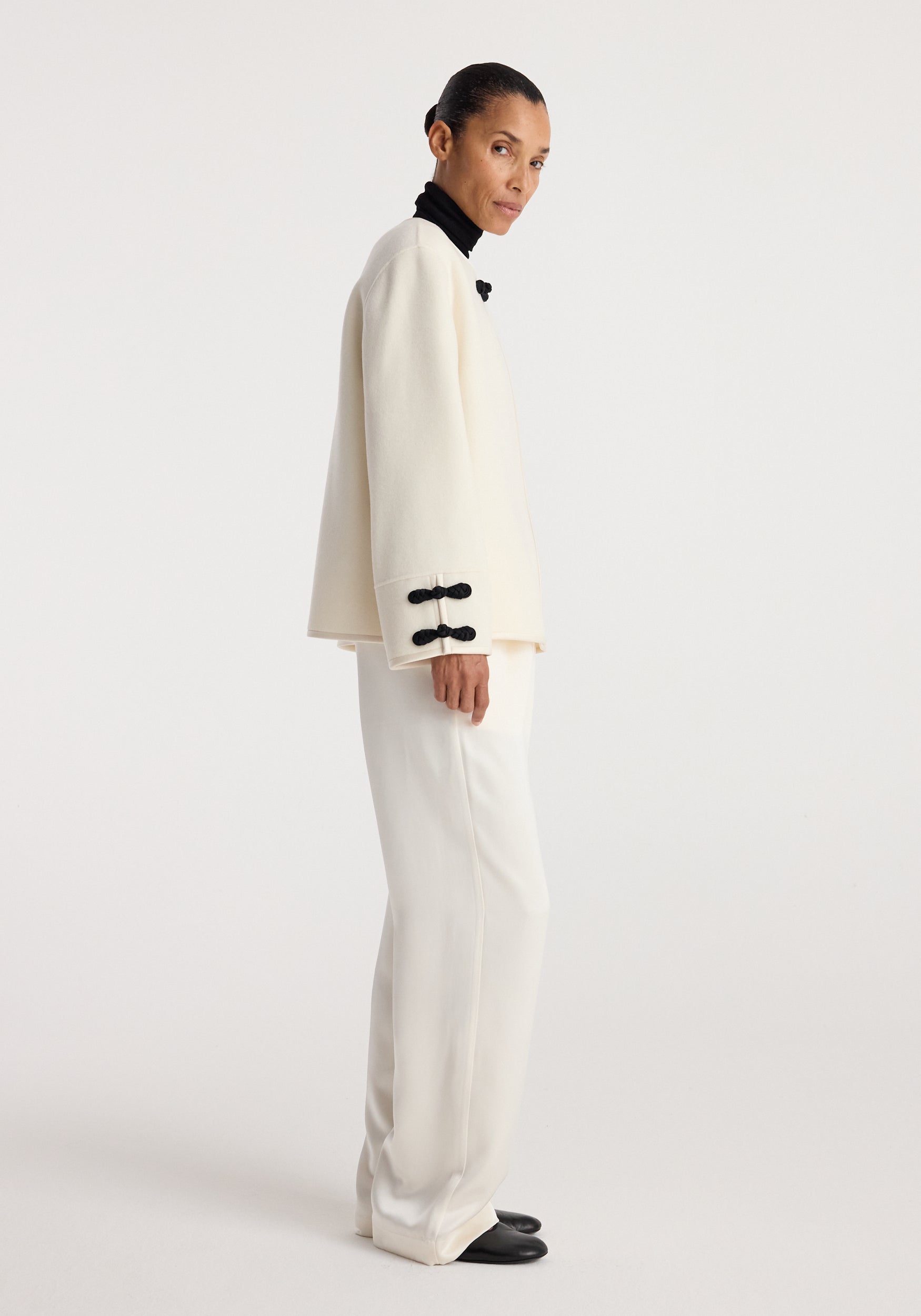 A model wearing a Double-faced pankou jacket | off white with black pankou button details on the cuffs and collar, paired with matching off-white pants and black shoes.