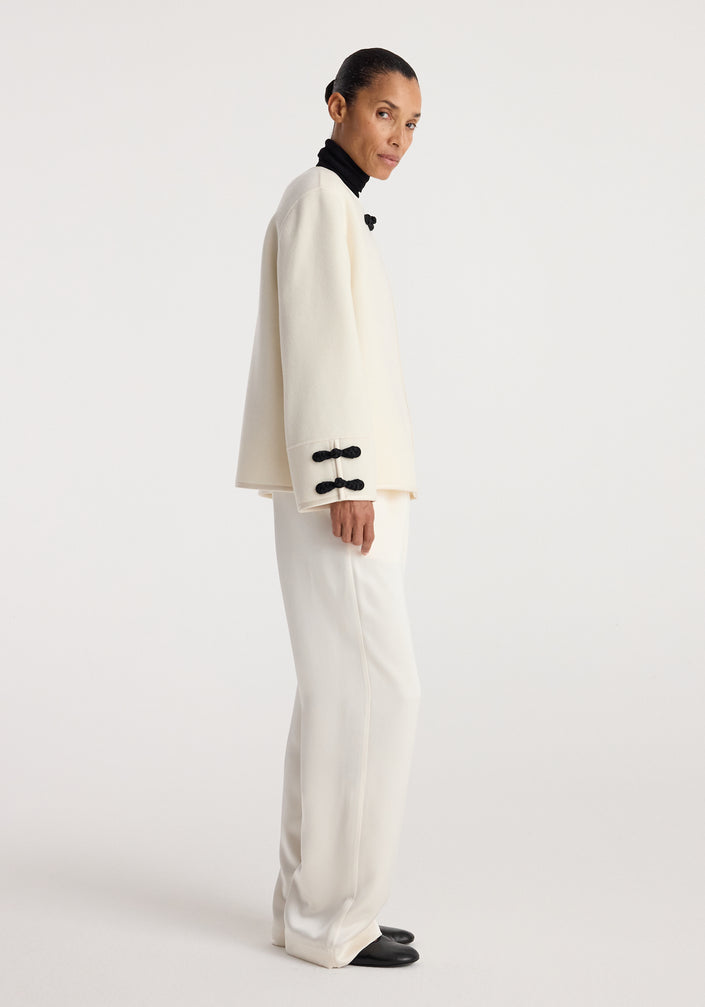 Woman wearing an off white Double-faced pankou jacket | off white with black decorative clasps, paired with matching wide-leg pants and black shoes against a plain background.