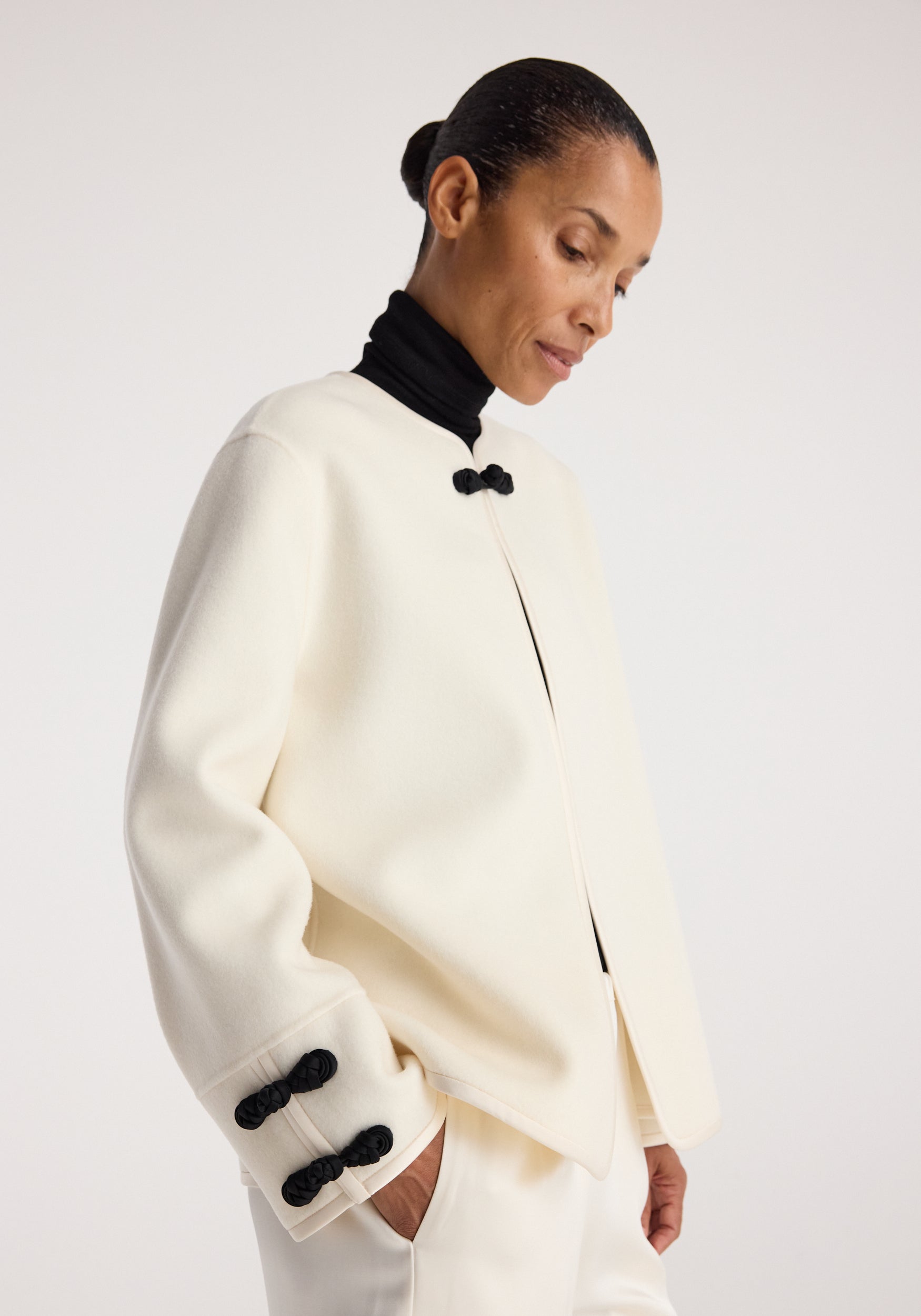 Woman wearing a stylish off white Double-faced pankou jacket | off white with black pankou knot closures on the collar and sleeves, paired with a black turtleneck and white pants.