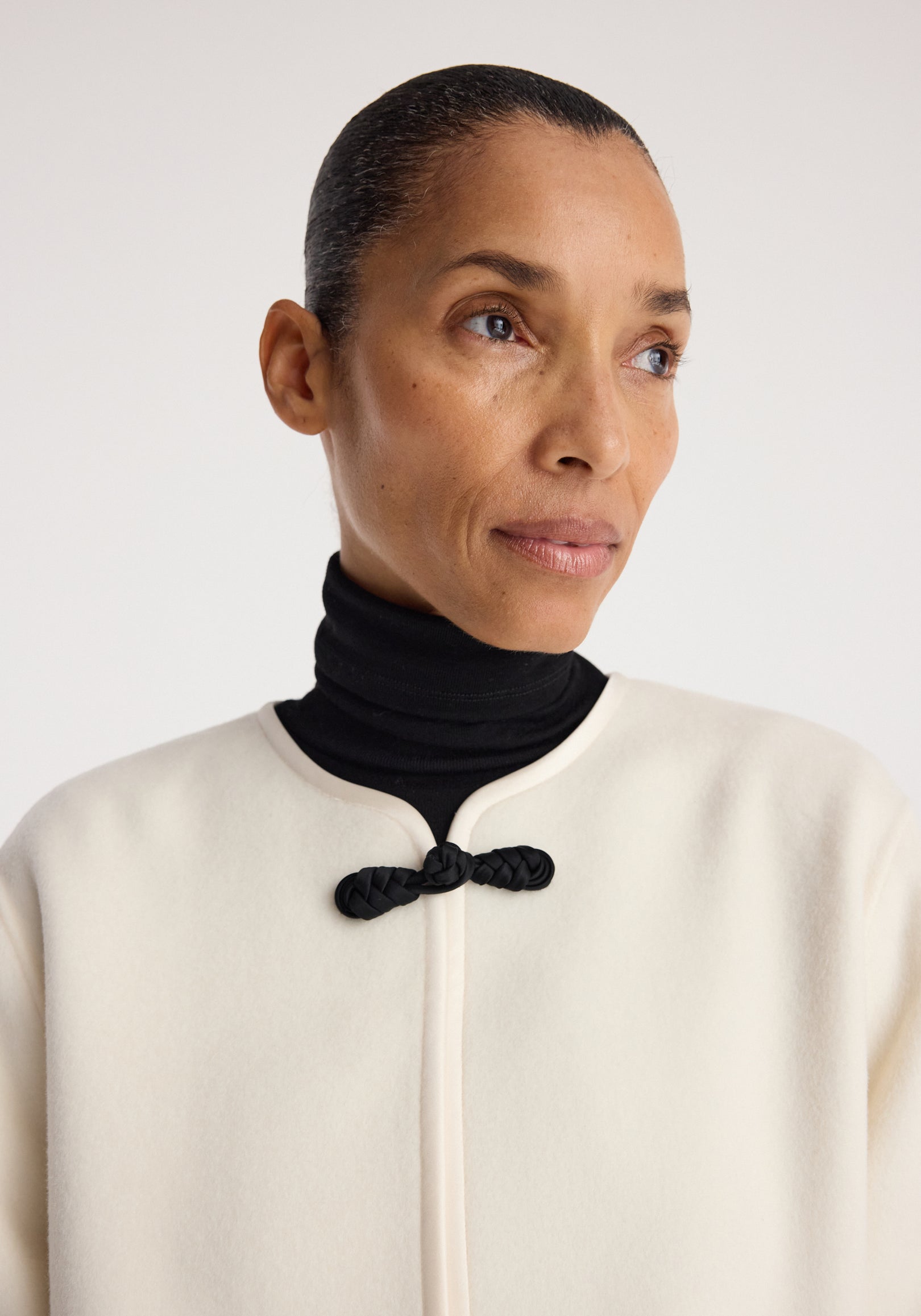 Close-up of a woman wearing a Double-faced pankou jacket | off white with a black pankou knot closure over a black turtleneck against a plain background.