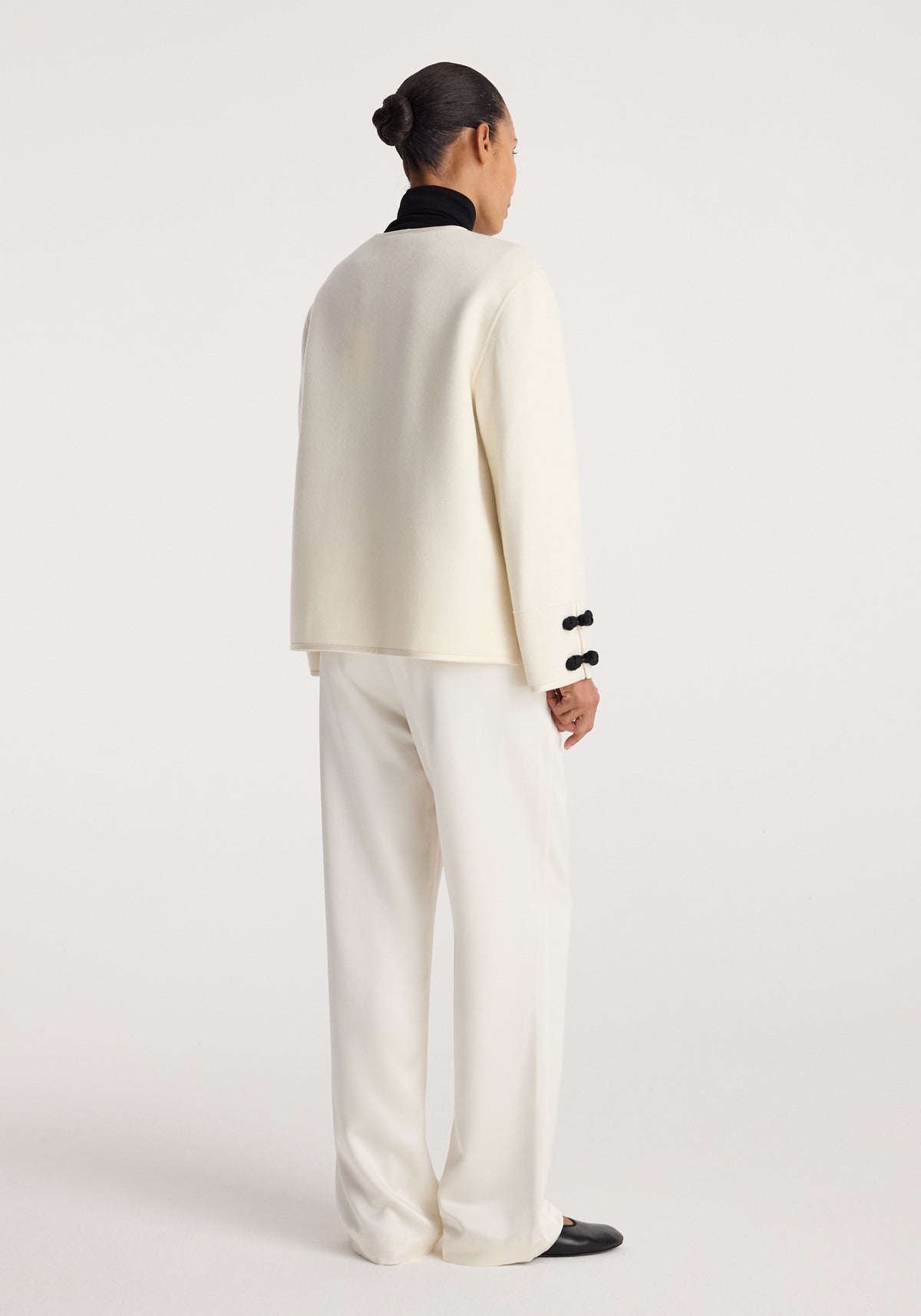 Back view of a person wearing a Double-faced pankou jacket | off white with black pankou button details on the cuffs, paired with loose off-white pants and black shoes.