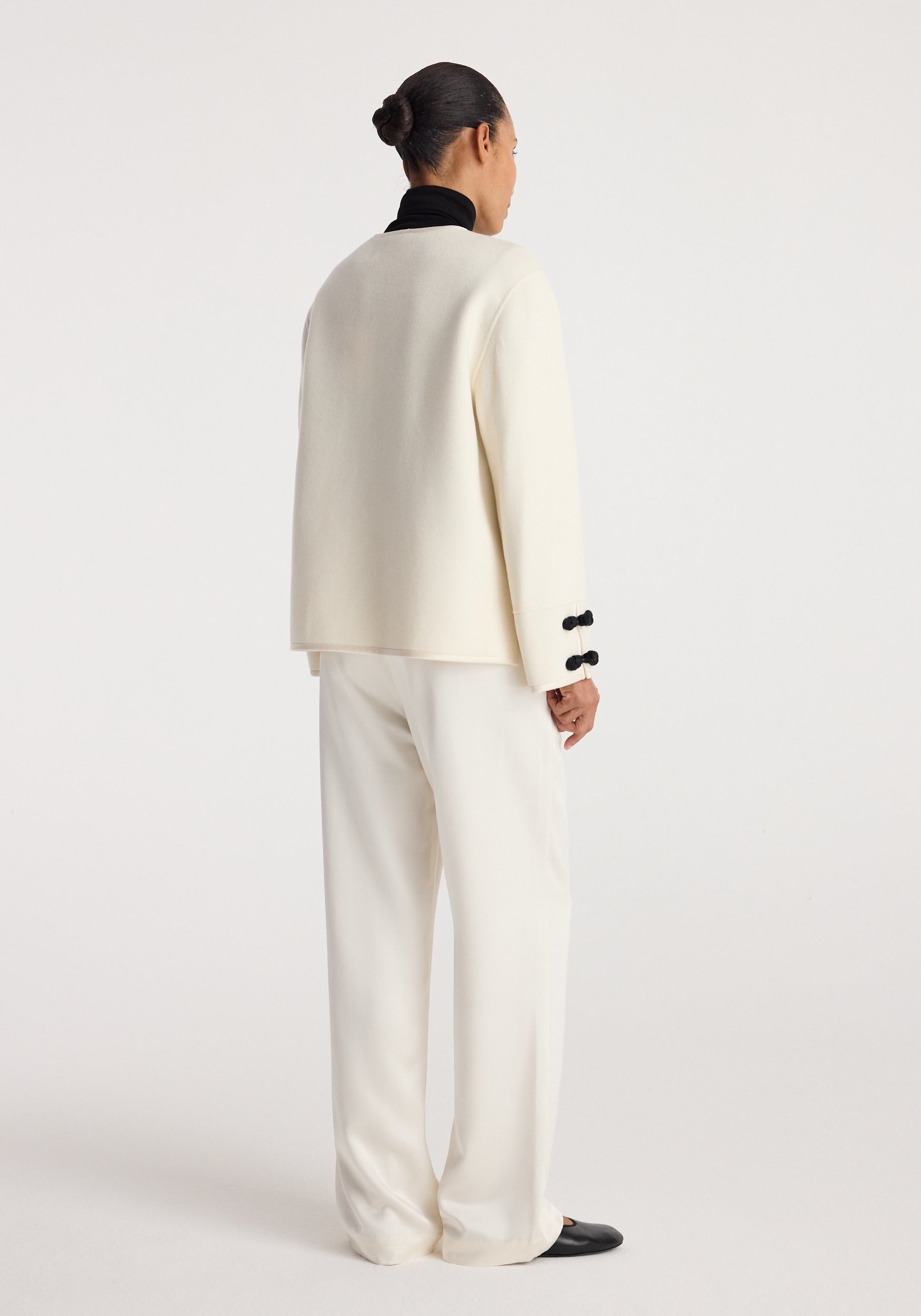 Back view of a person wearing a Double-faced pankou jacket | off white with black pankou button details on the cuffs, paired with loose off-white pants and black shoes.