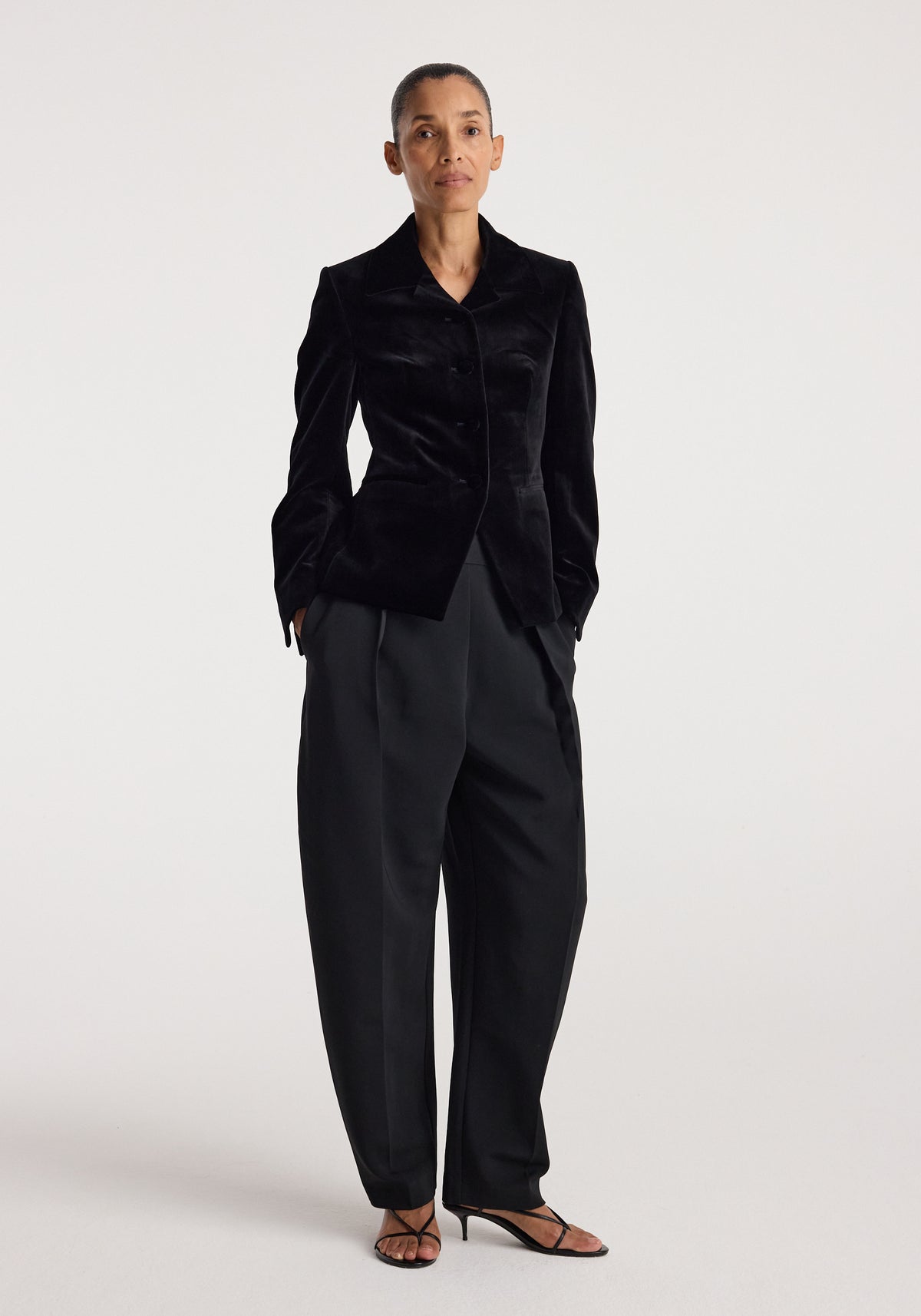 Woman wearing a classic fitted velvet blazer | black styled with wide-leg black trousers and strappy heels, standing against a plain white background.