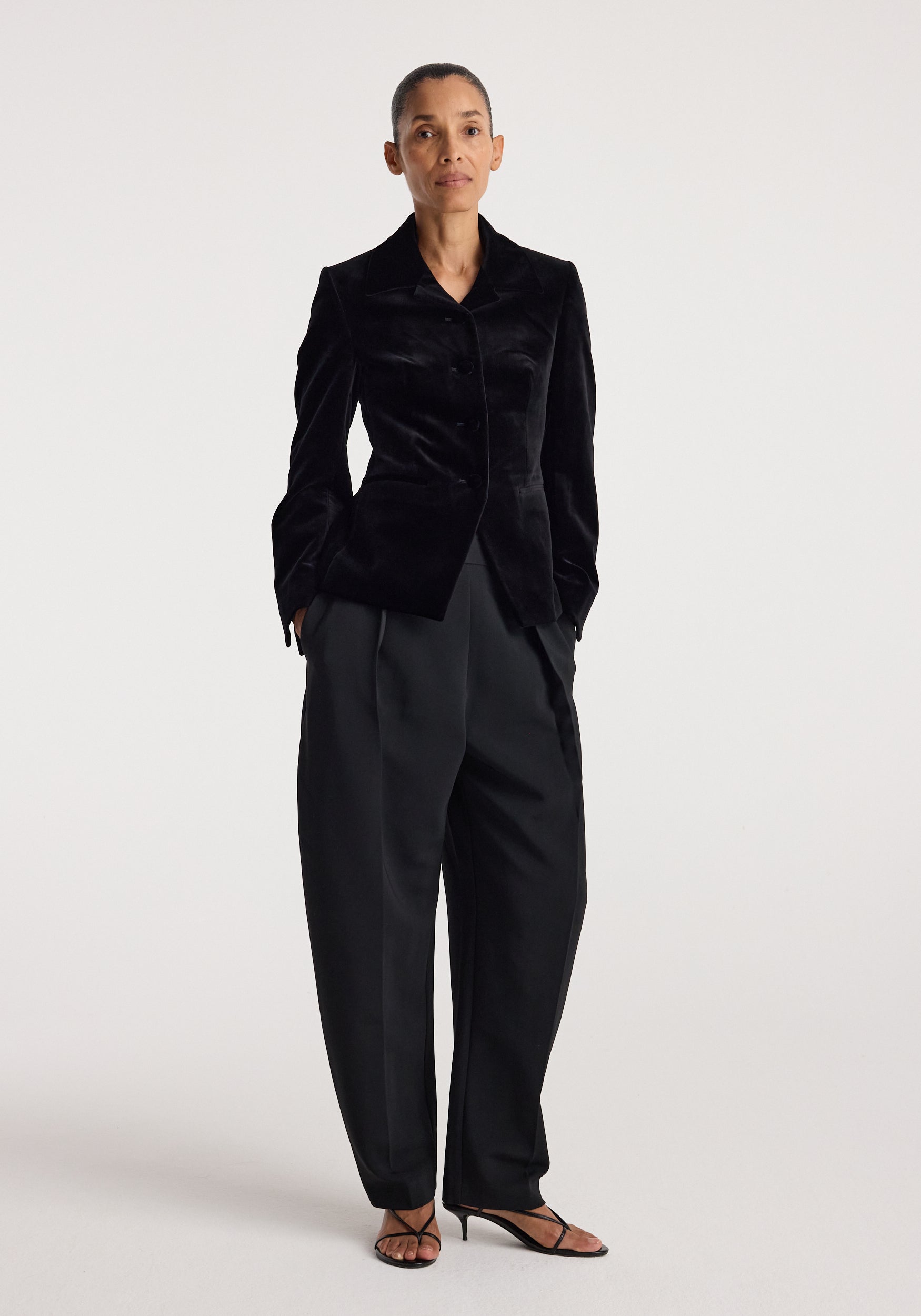 Woman wearing a classic fitted velvet blazer | black styled with wide-leg black trousers and strappy heels, standing against a plain white background.