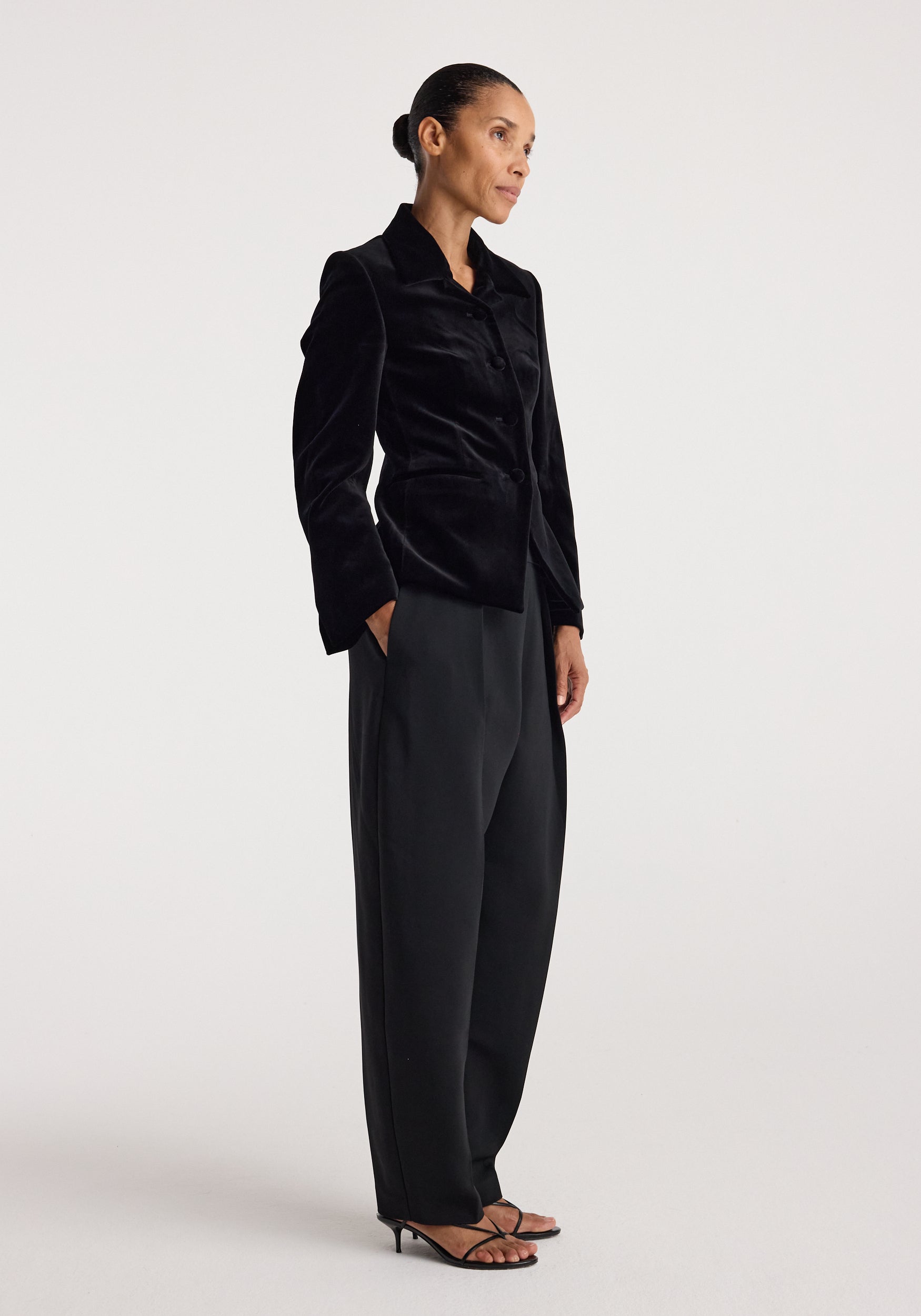 Woman wearing a Classic fitted velvet blazer | black with a tailored silhouette, paired with loose black trousers and strappy heels in a minimalist studio setting.
