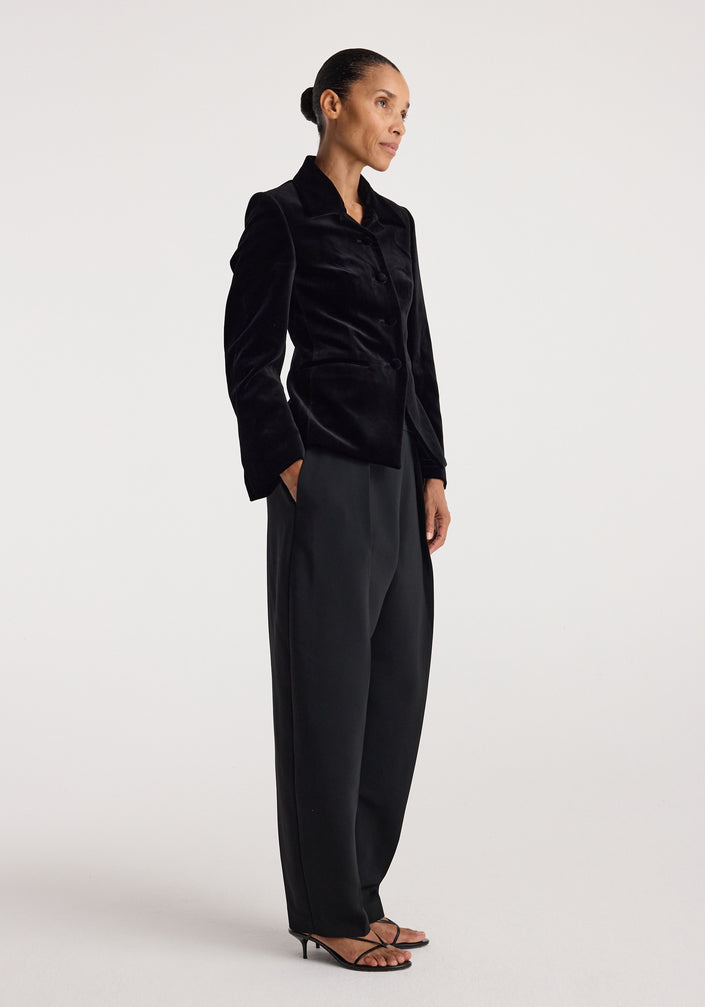 Woman wearing a classic fitted velvet blazer | black styled with wide-leg black trousers and strappy heels, standing against a plain white background.