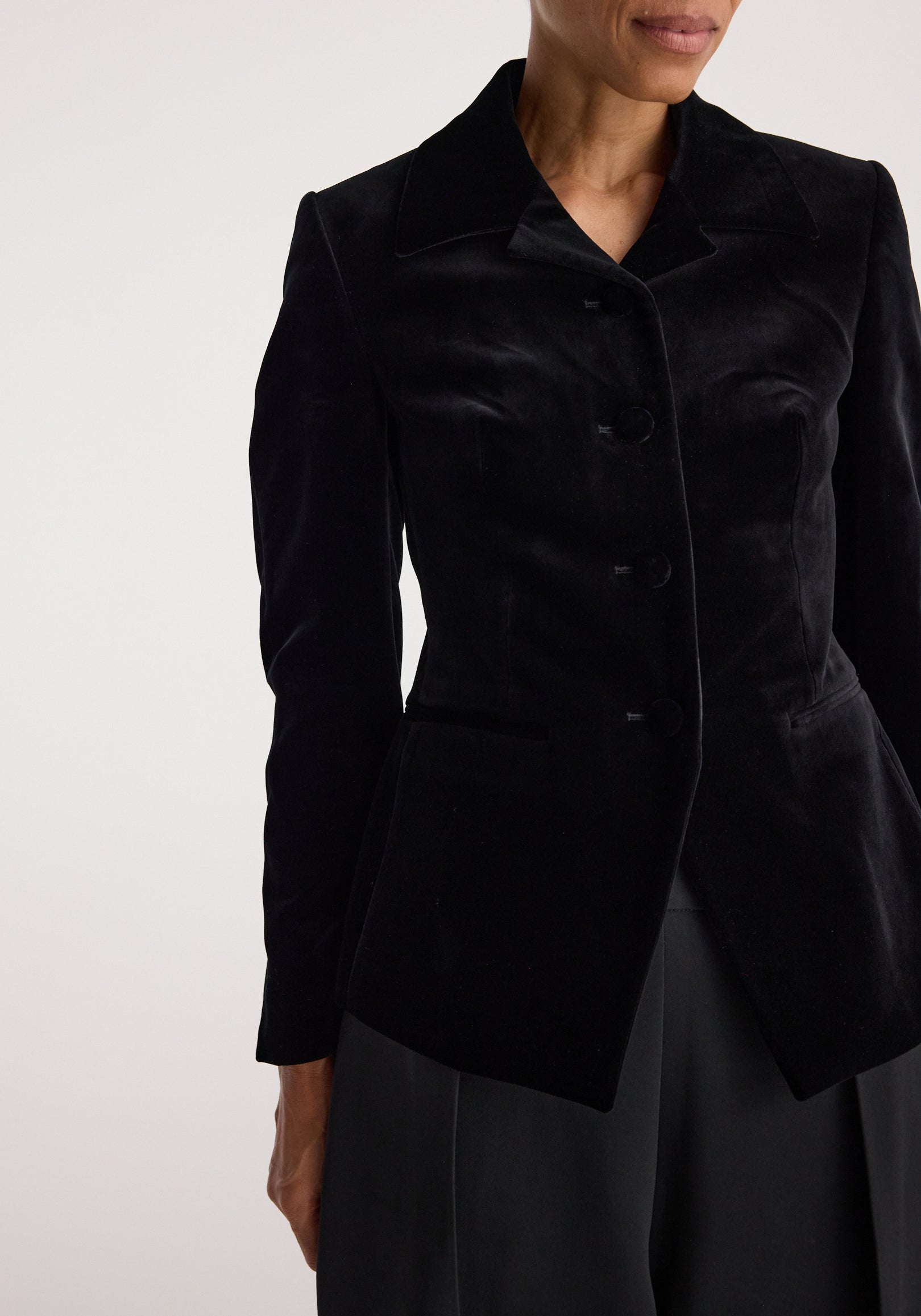 Close-up of a person wearing a Classic fitted velvet blazer | black with a tailored collar and buttons down the front, styled with black trousers.