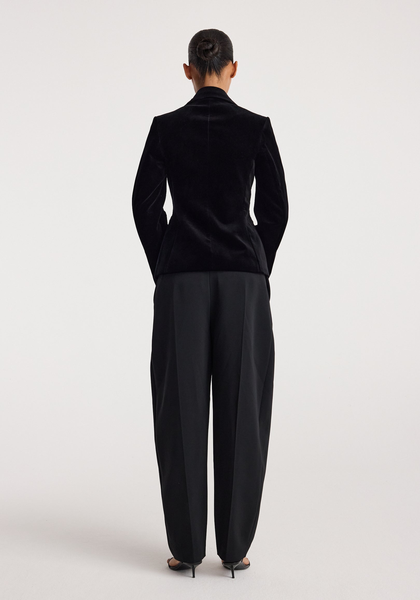Woman wearing classic fitted velvet blazer | black viewed from the back, paired with black wide-leg trousers and black heels, standing against a plain light background.