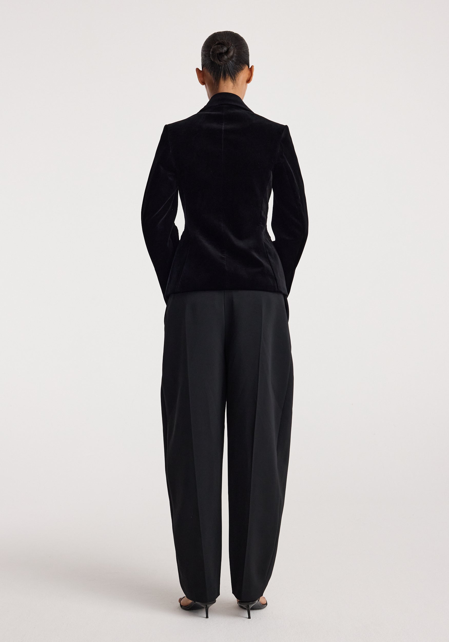 Woman wearing classic fitted velvet blazer | black viewed from the back, paired with black wide-leg trousers and black heels, standing against a plain light background.