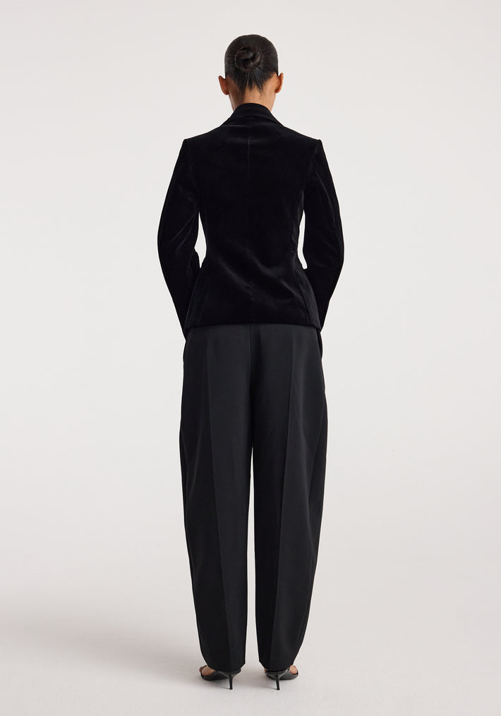 Woman wearing classic fitted velvet blazer | black viewed from the back, paired with black wide-leg trousers and black heels, standing against a plain light background.