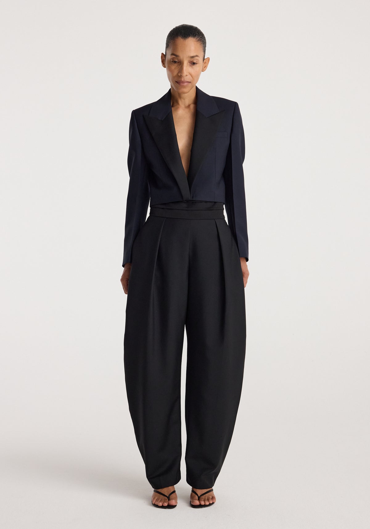 Woman wearing a tailored Cropped blazer | navy with a deep neckline paired with high-waisted wide-leg black trousers and strappy black sandals against a plain background