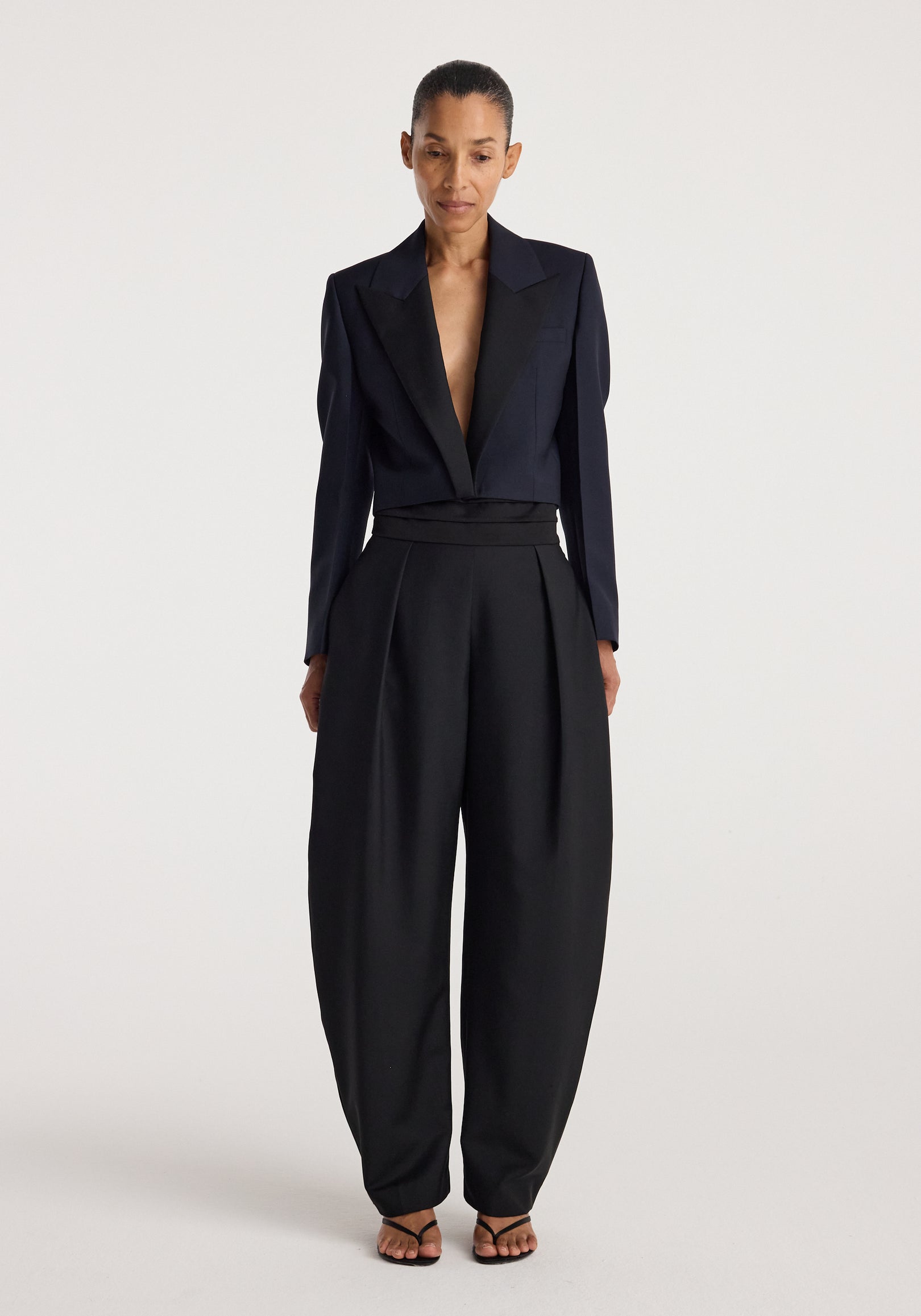 Woman wearing a tailored Cropped blazer | navy with a deep neckline paired with high-waisted wide-leg black trousers and strappy black sandals against a plain background