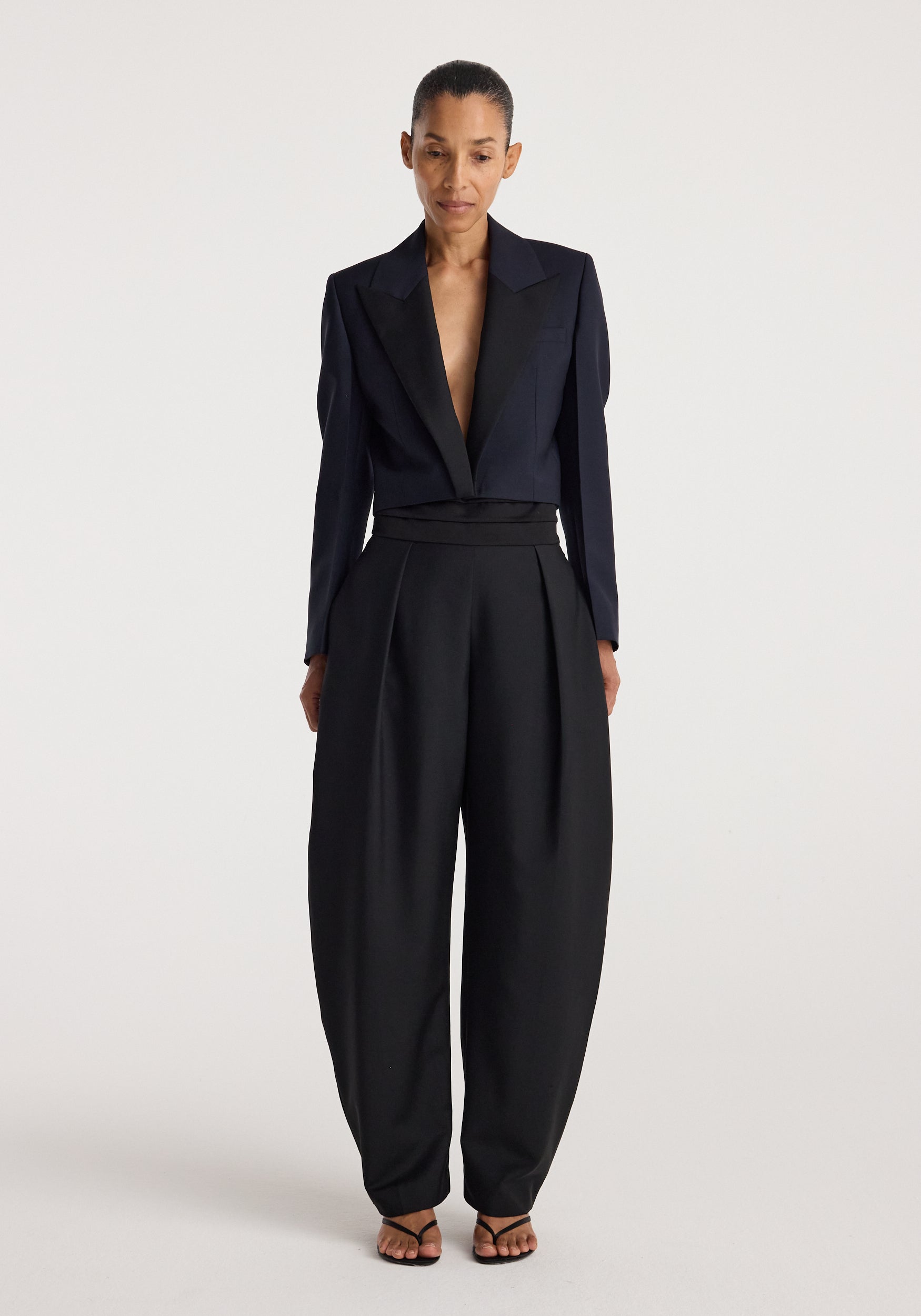 Woman wearing a tailored Cropped blazer | navy with a deep neckline paired with high-waisted wide-leg black trousers and strappy black sandals against a plain background
