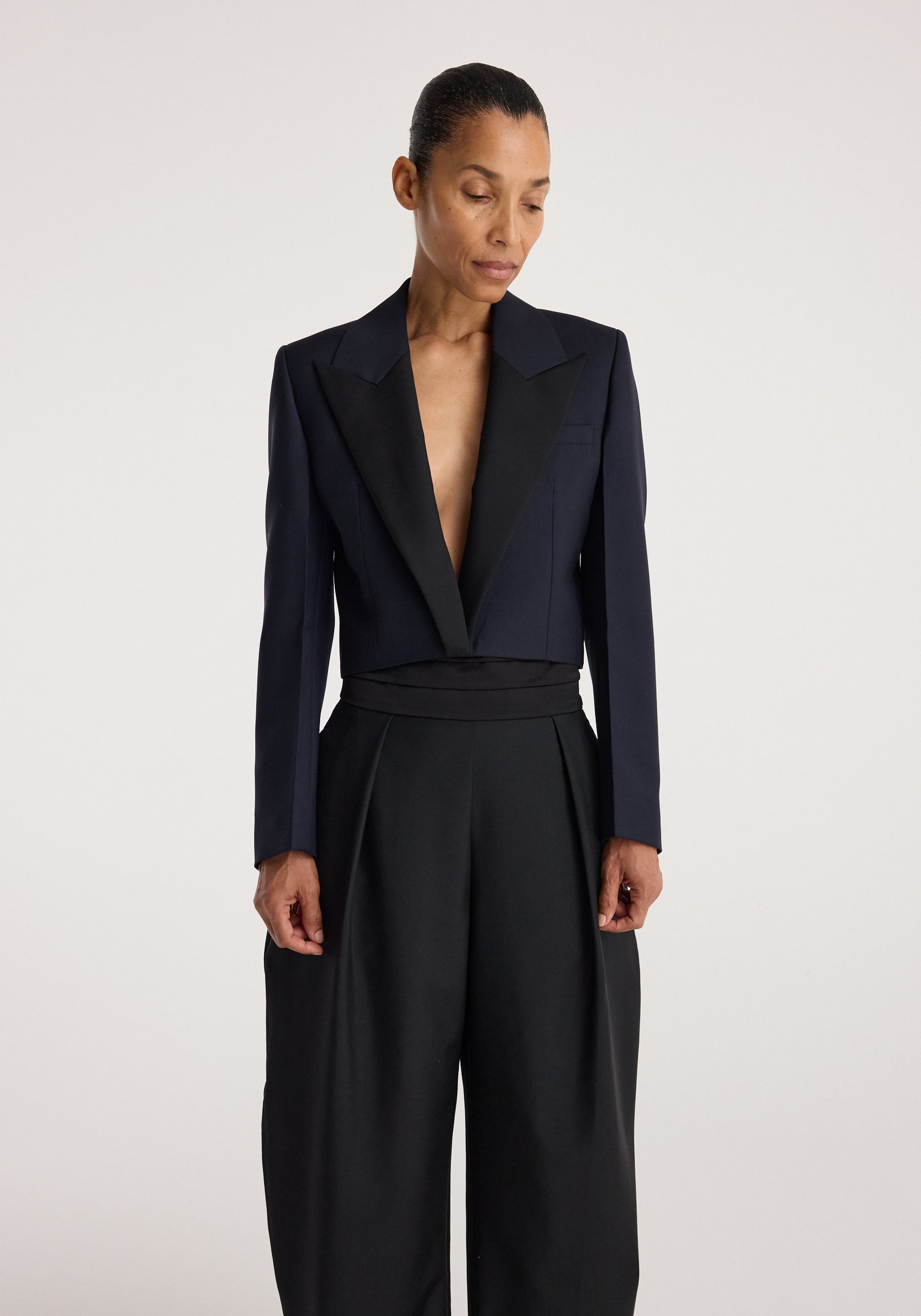 Woman wearing a sleek cropped blazer | navy with contrasting lapels, paired with high-waisted black wide-leg pants, standing against a plain light background.