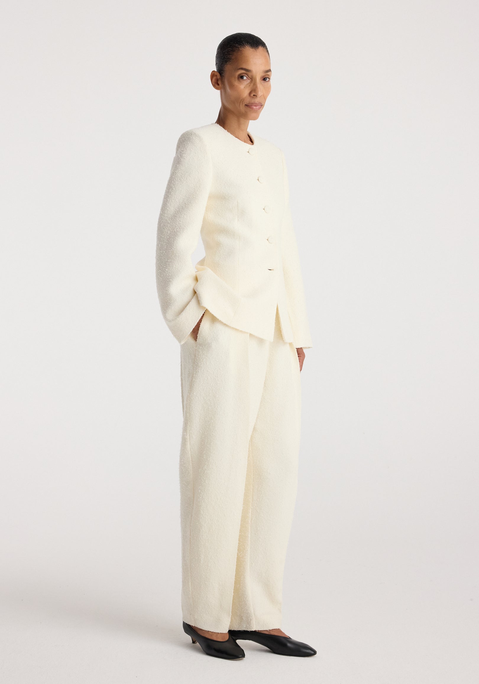 Woman wearing a cream textured Bouclé shaped blazer | cream with matching wide-leg pants and black pointed-toe shoes, standing against a plain light background.