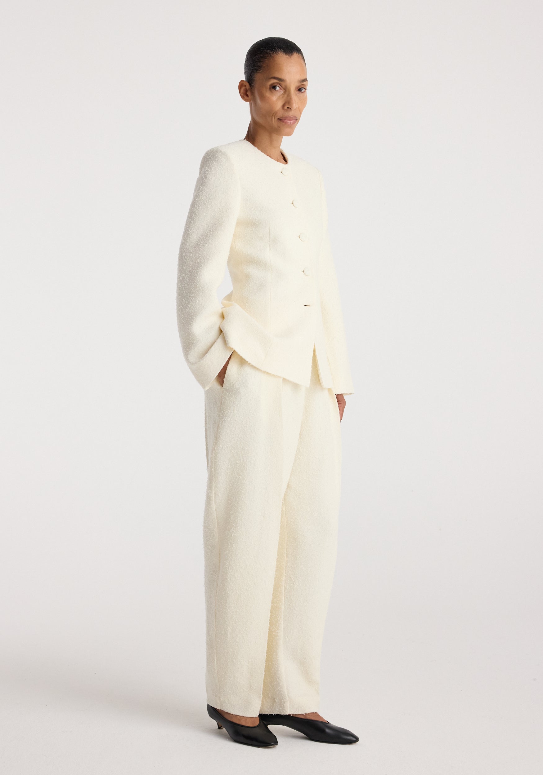 Woman wearing a cream textured Bouclé shaped blazer | cream with matching wide-leg pants and black pointed-toe shoes, standing against a plain light background.