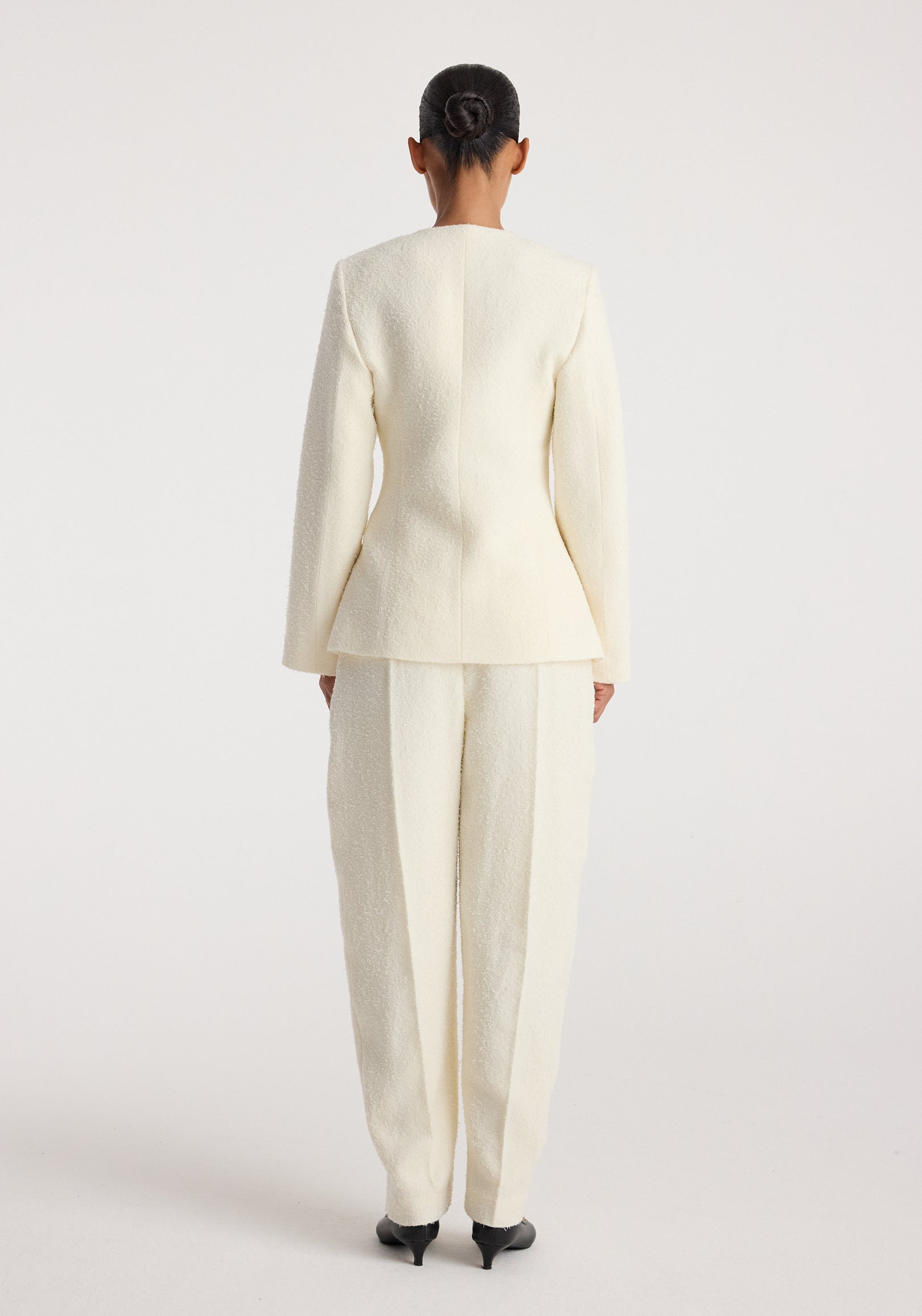 Back view of a person wearing a cream textured bouclé shaped blazer with matching cream trousers and black heeled shoes, styled with hair in a neat bun, showcasing the blazer's tailored fit and structure.