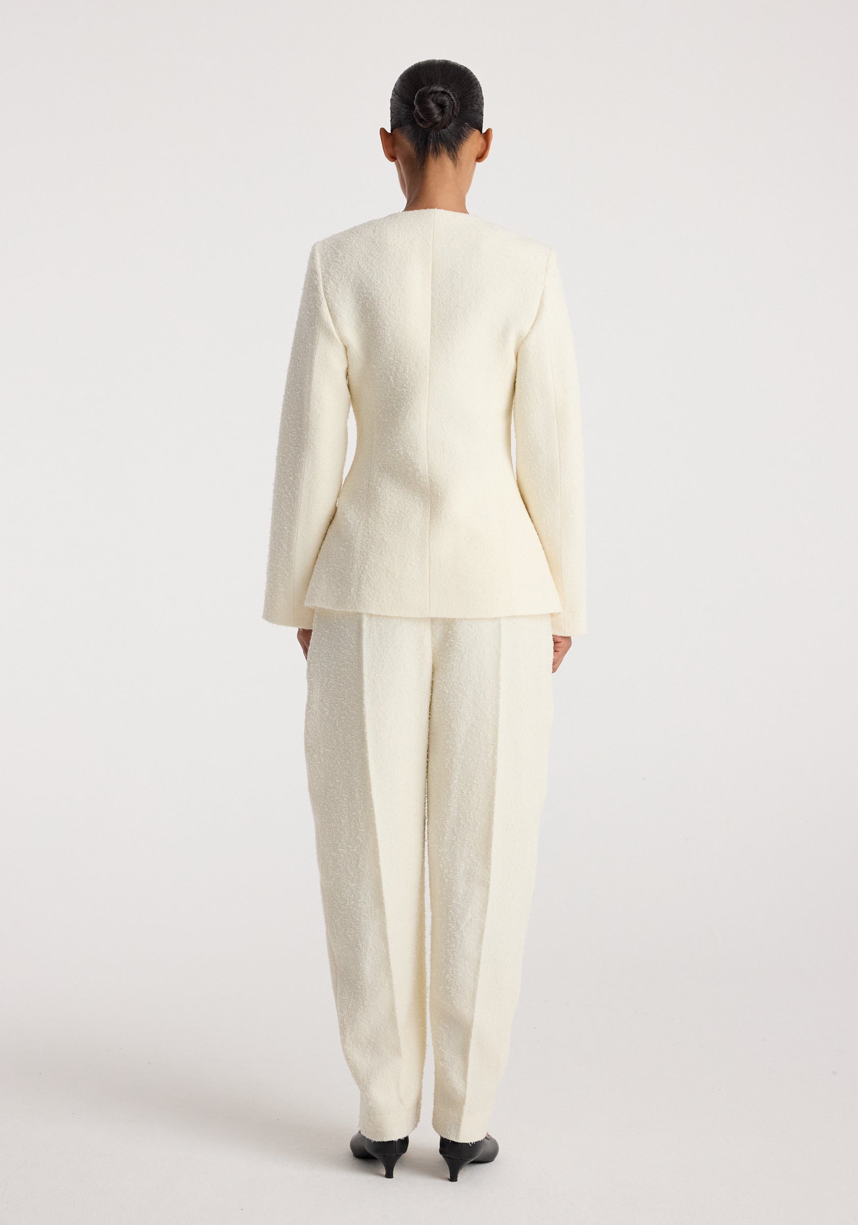 Back view of a person wearing a cream textured bouclé shaped blazer with matching cream trousers and black heeled shoes, styled with hair in a neat bun, showcasing the blazer's tailored fit and structure.