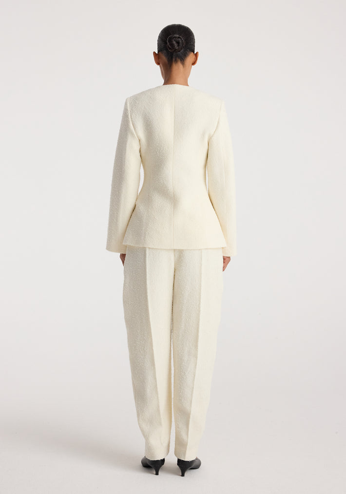 Back view of a person wearing a cream textured bouclé shaped blazer with matching cream trousers and black heeled shoes, styled with hair in a neat bun, showcasing the blazer's tailored fit and structure.