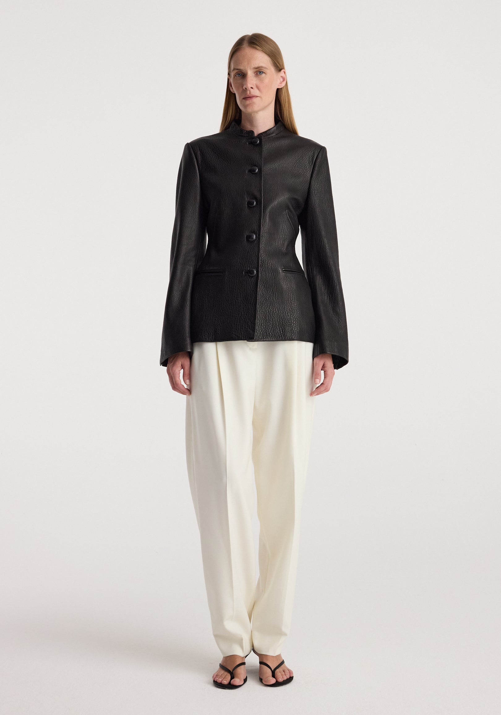 Woman wearing a fitted black shaped grain leather jacket | black with button closure, paired with loose white trousers and black open-toed sandals, standing against a plain background.