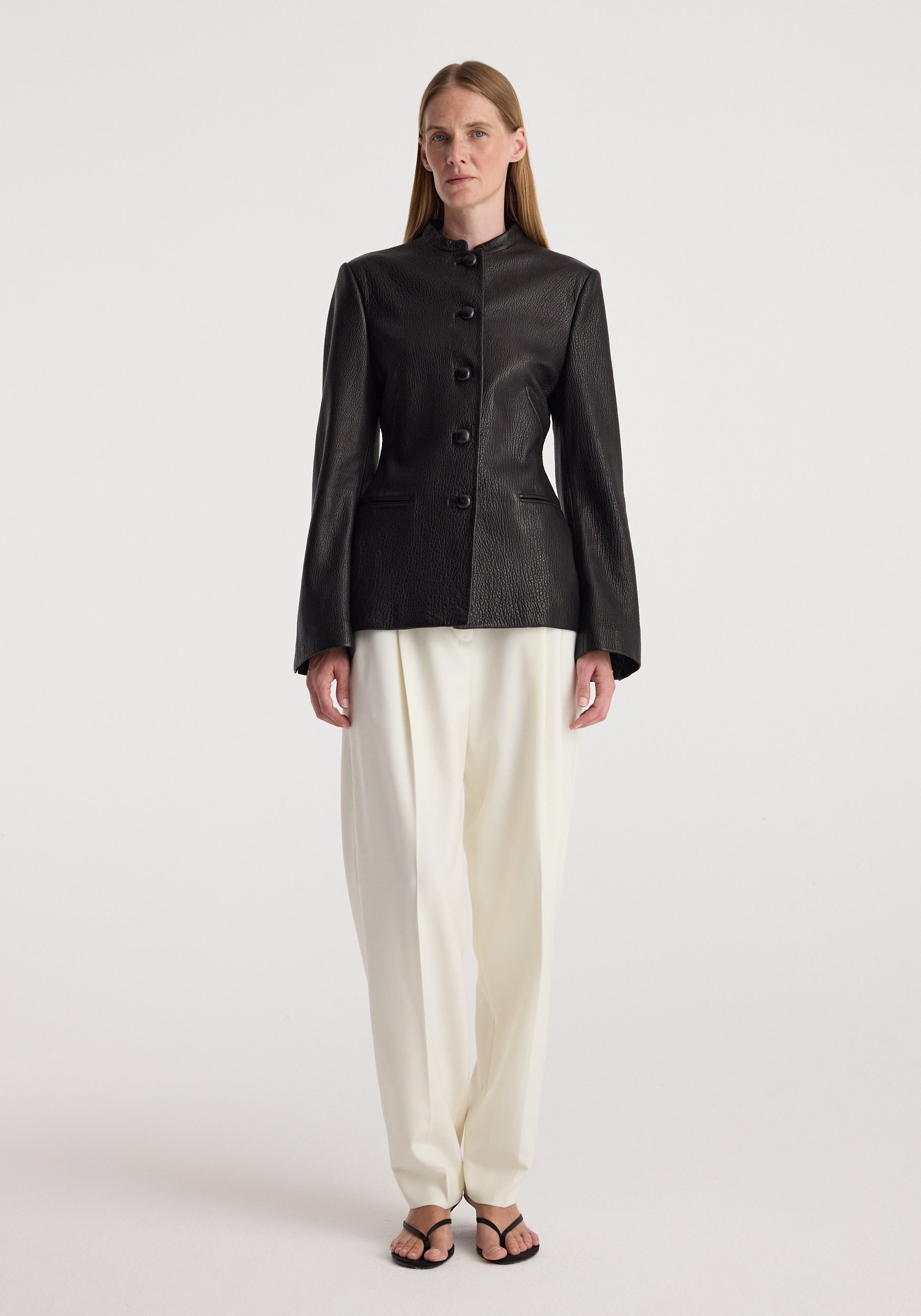 Woman wearing a fitted black shaped grain leather jacket | black with button closure, paired with loose white trousers and black open-toed sandals, standing against a plain background.