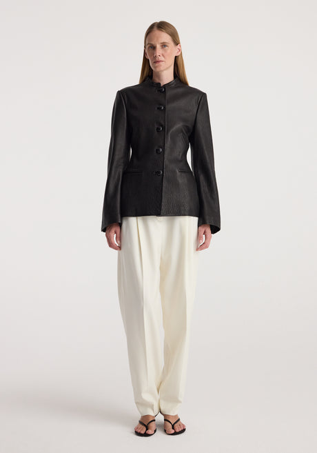 Woman wearing a fitted black shaped grain leather jacket | black with button closure, paired with loose white trousers and black open-toed sandals, standing against a plain background.