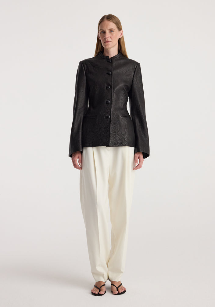 Woman wearing a fitted black shaped grain leather jacket | black with button closure, paired with loose white trousers and black open-toed sandals, standing against a plain background.