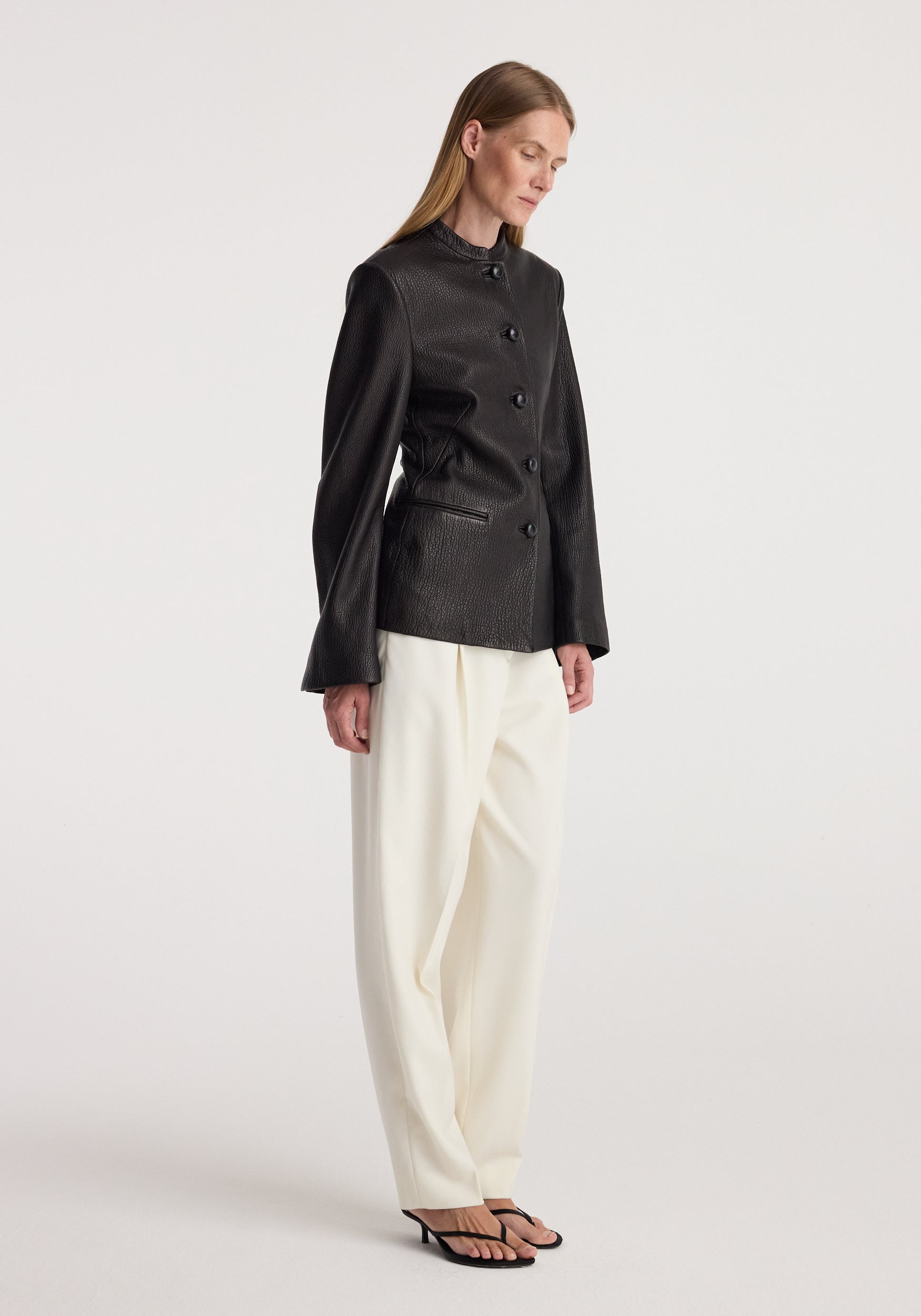 Woman wearing a sleek black shaped grain leather jacket | black with long sleeves and button closure, paired with loose white trousers and black heeled sandals, standing sideways.