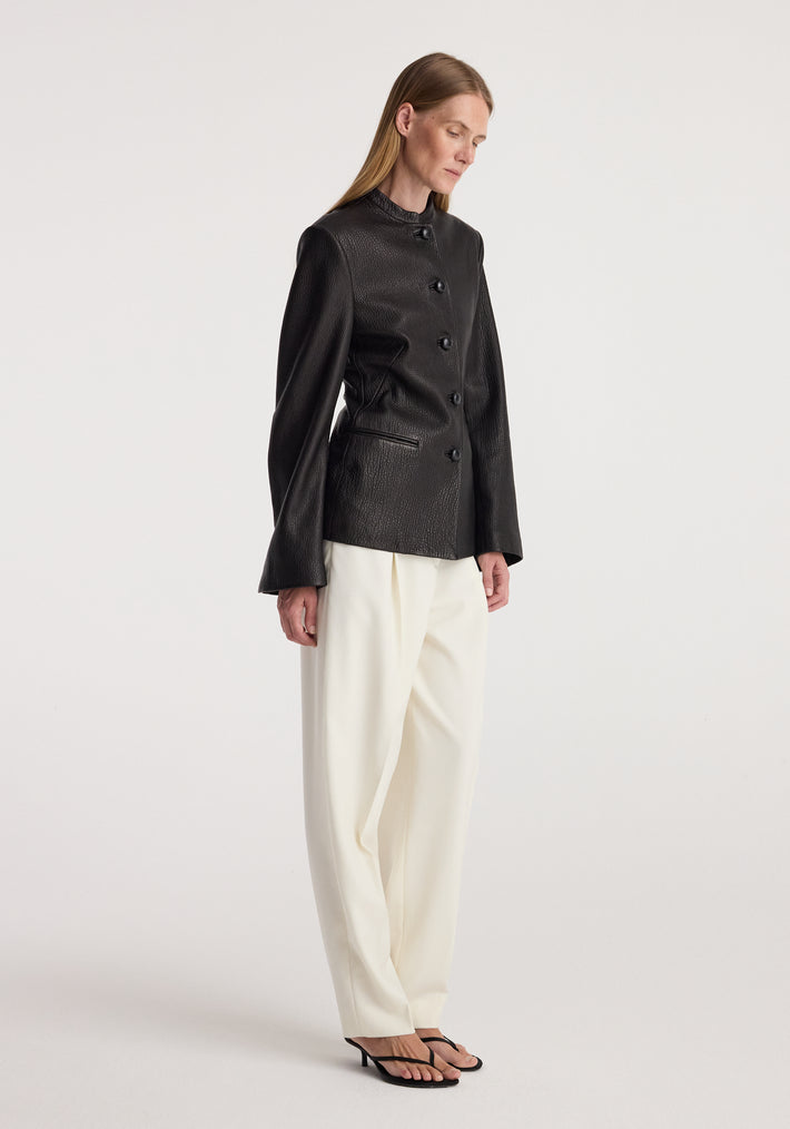 Woman wearing a sleek black shaped grain leather jacket | black with long sleeves and button closure, paired with loose white trousers and black heeled sandals, standing sideways.