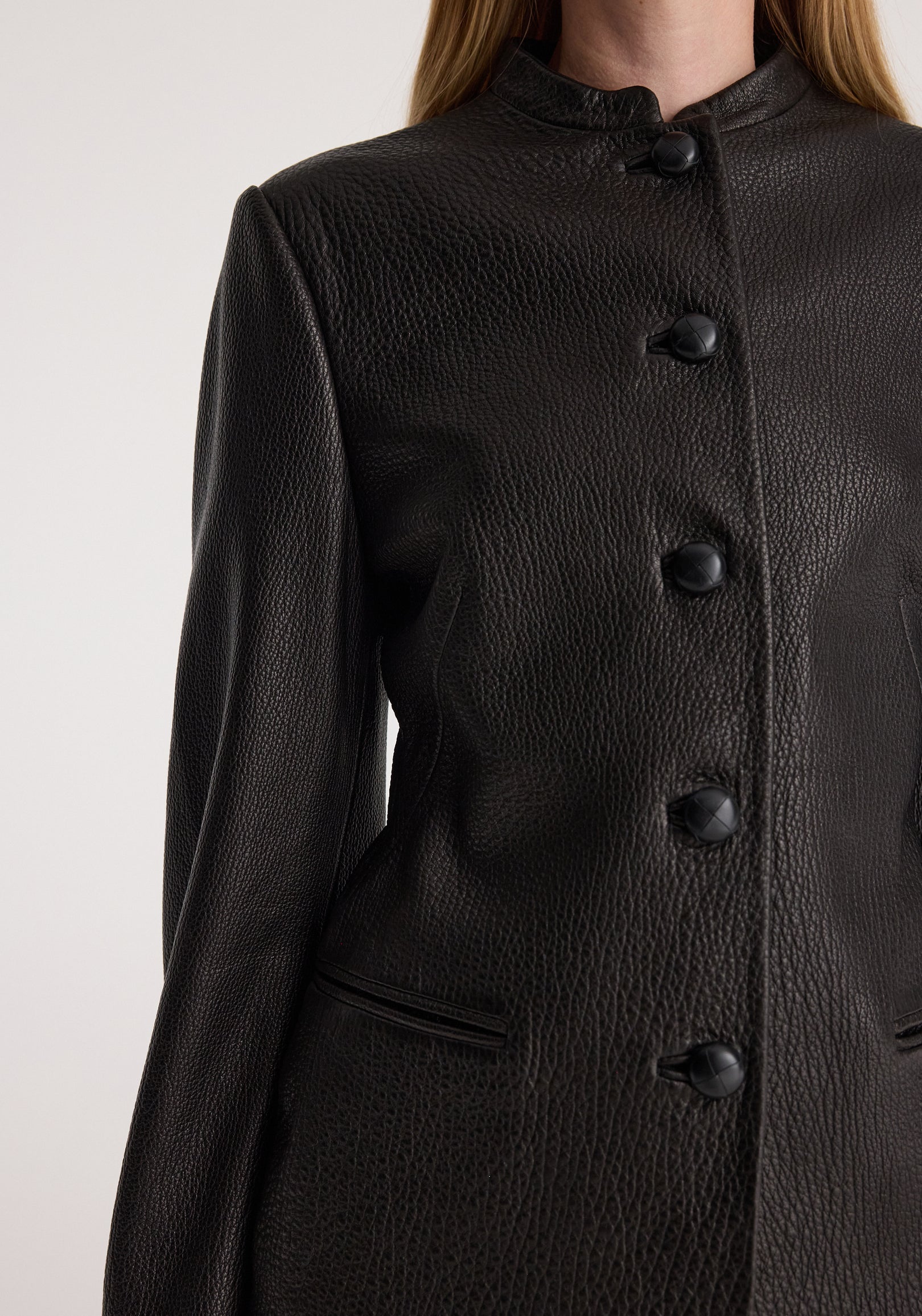 Close-up of a woman wearing a shaped grain leather jacket | black, showcasing the textured leather with a high collar and large button closures down the front.