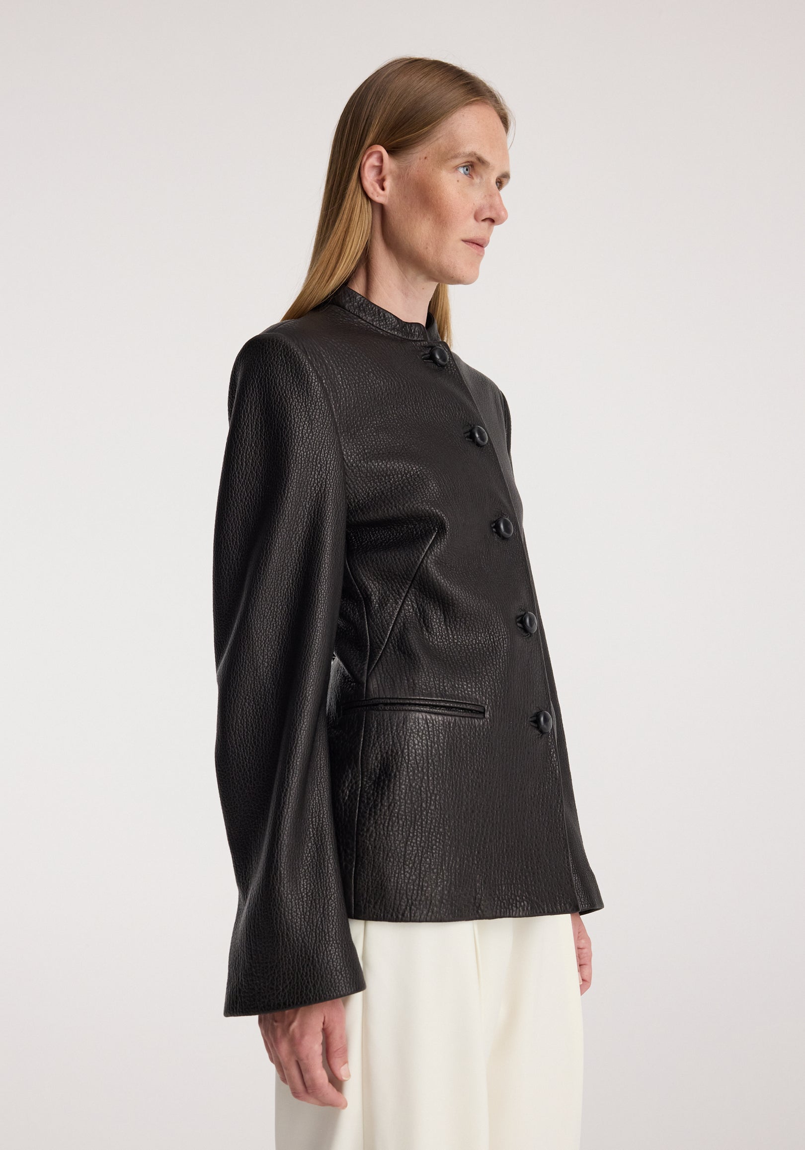 Woman wearing a black Shaped grain leather jacket | black with textured finish, buttoned front, high collar, and long sleeves, paired with white pants against a plain background.