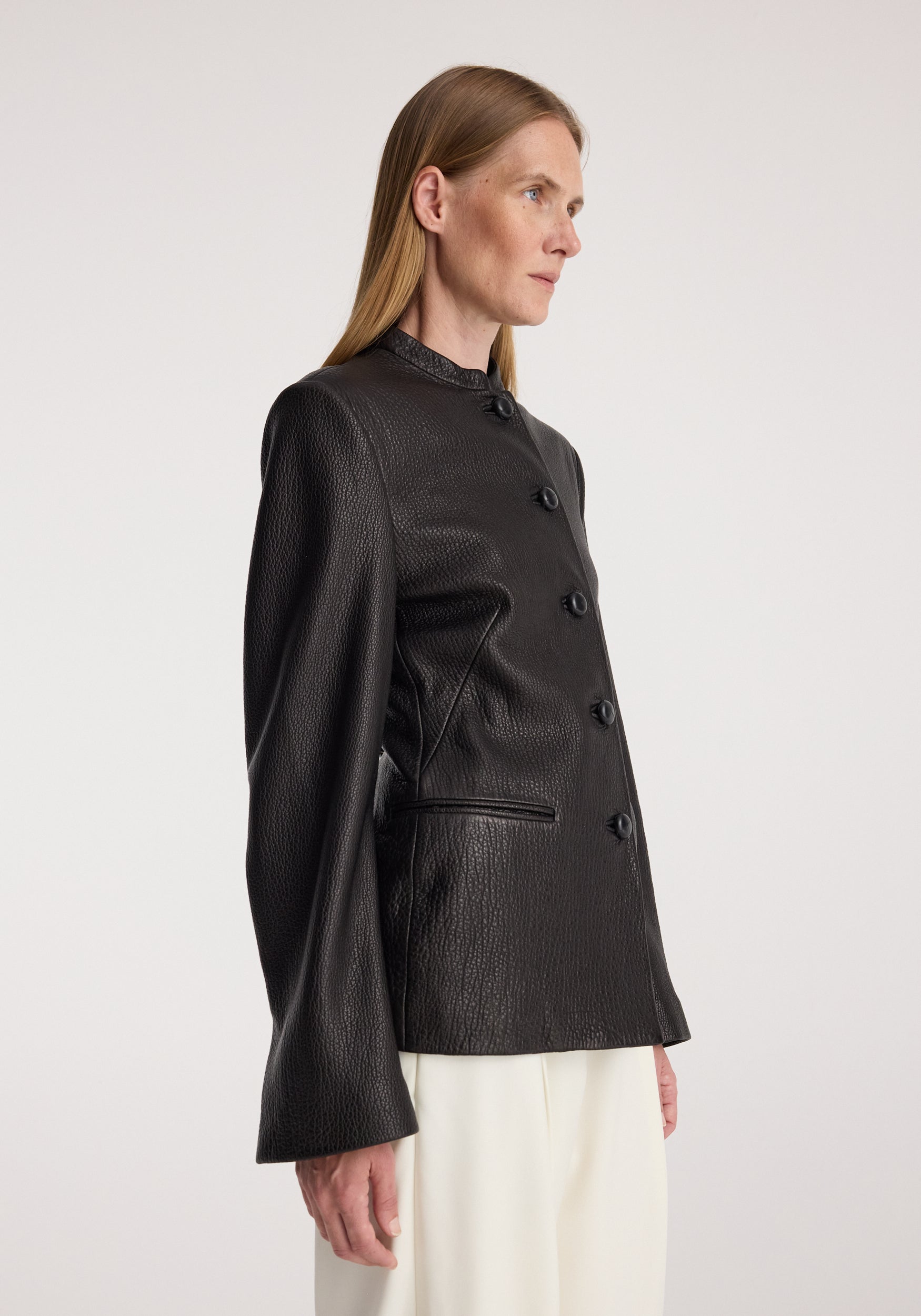 Woman wearing a black Shaped grain leather jacket | black with textured finish, buttoned front, high collar, and long sleeves, paired with white pants against a plain background.