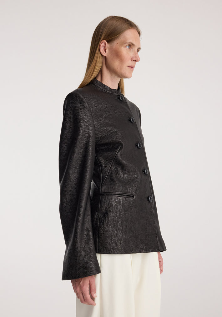 Woman wearing a black Shaped grain leather jacket | black with textured finish, buttoned front, high collar, and long sleeves, paired with white pants against a plain background.