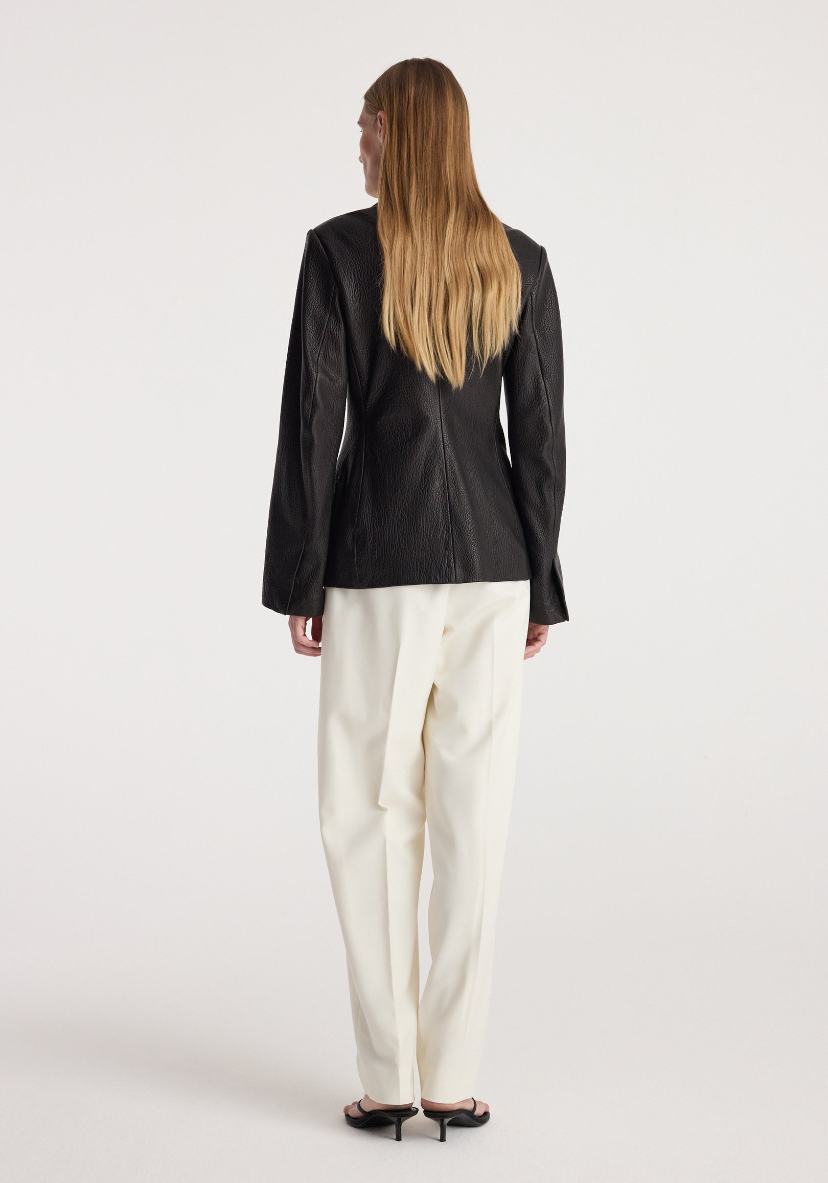 Woman seen from the back wearing a Shaped grain leather jacket | black paired with white trousers and black heeled sandals against a plain background.