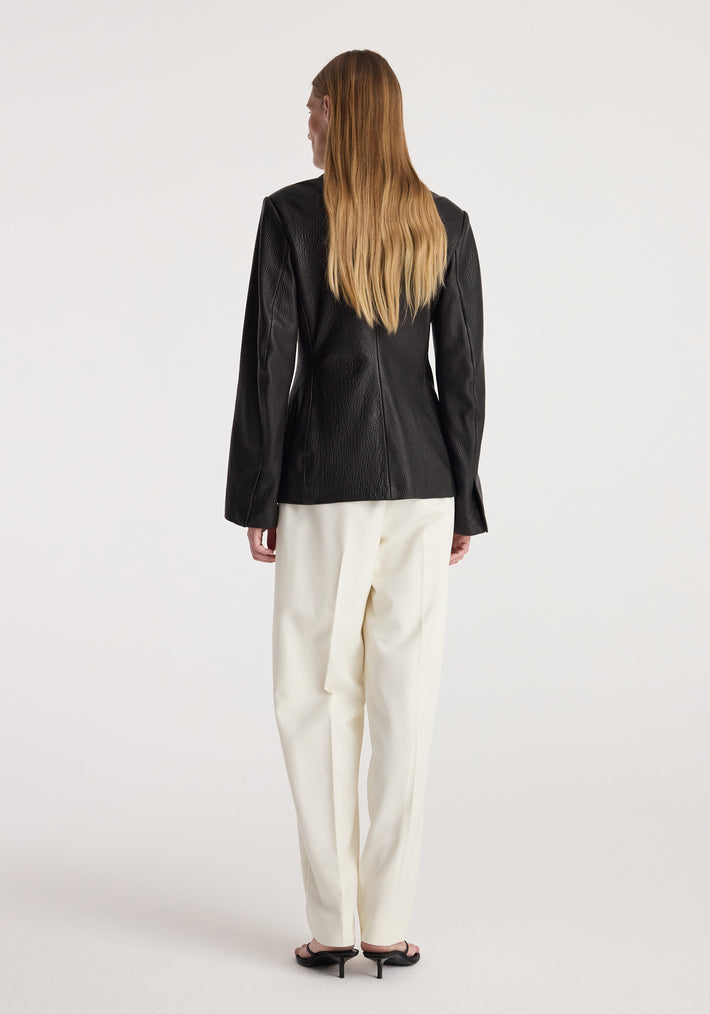 Woman seen from the back wearing a Shaped grain leather jacket | black paired with white trousers and black heeled sandals against a plain background.