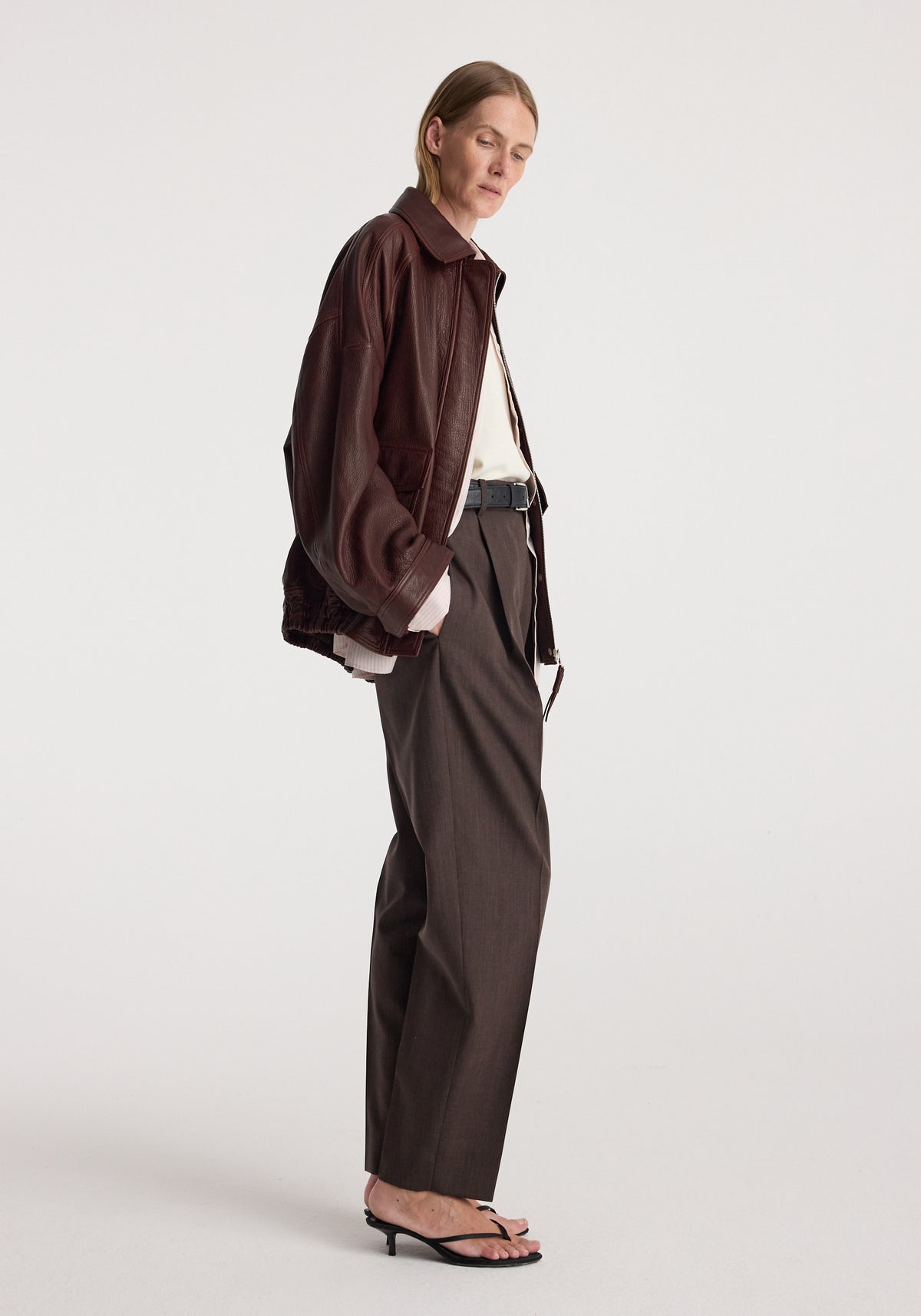 Woman wearing a grain leather aviator jacket | chestnut in a relaxed fit paired with high-waisted dark trousers and black heeled sandals against a plain background.
