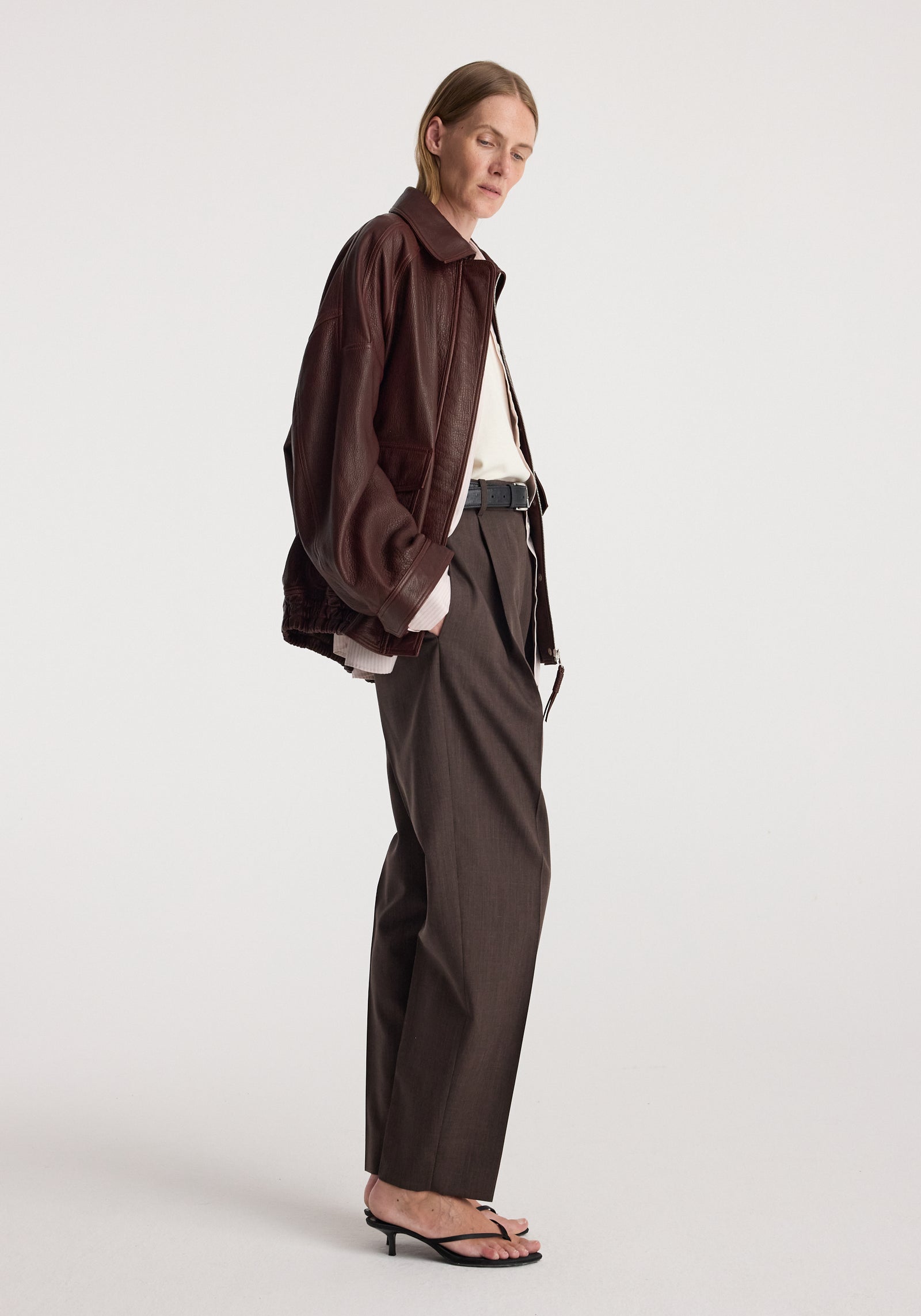 Woman wearing a grain leather aviator jacket | chestnut in a relaxed fit paired with high-waisted dark trousers and black heeled sandals against a plain background.
