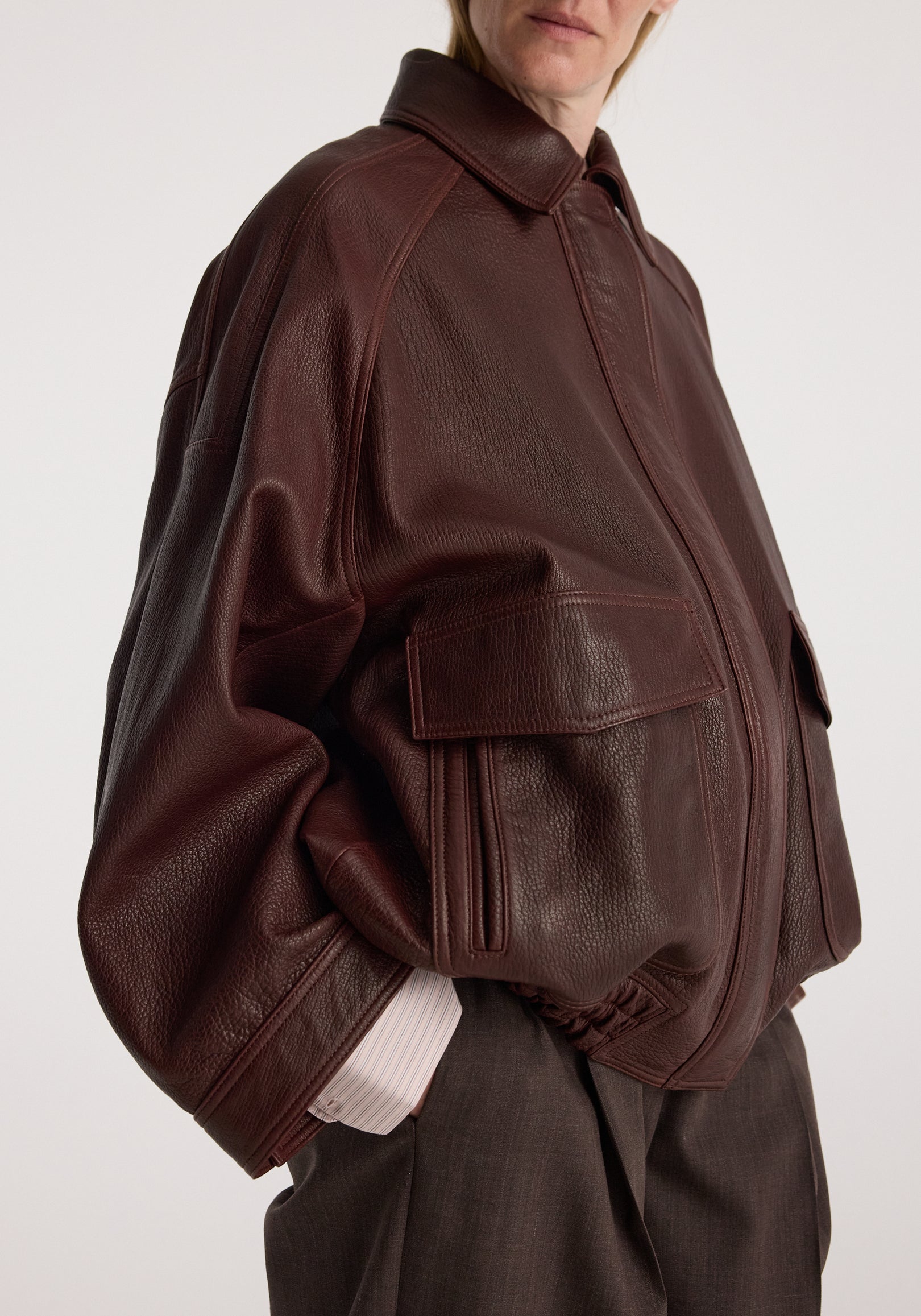 Close-up of a person wearing a chestnut Grain leather aviator jacket with large front pockets and a spread collar, styled with dark pants and a light shirt cuff visible.